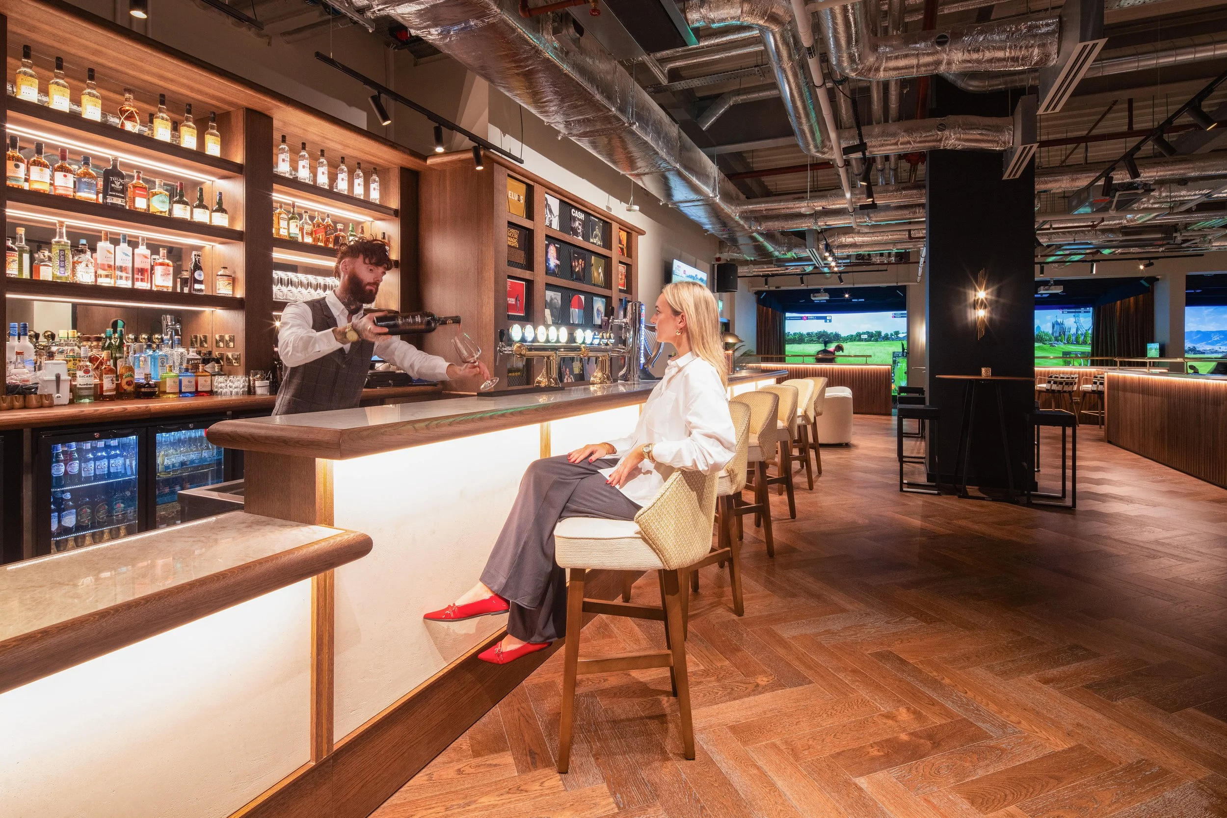 A woman sitting at a bar counter in a modern, upscale bar or lounge, with a bartender pouring her a drink. The bar has bottles of alcohol on shelves behind it, and the room features wood flooring, industrial-style ceiling pipes, and large windows showing a green landscape outside.