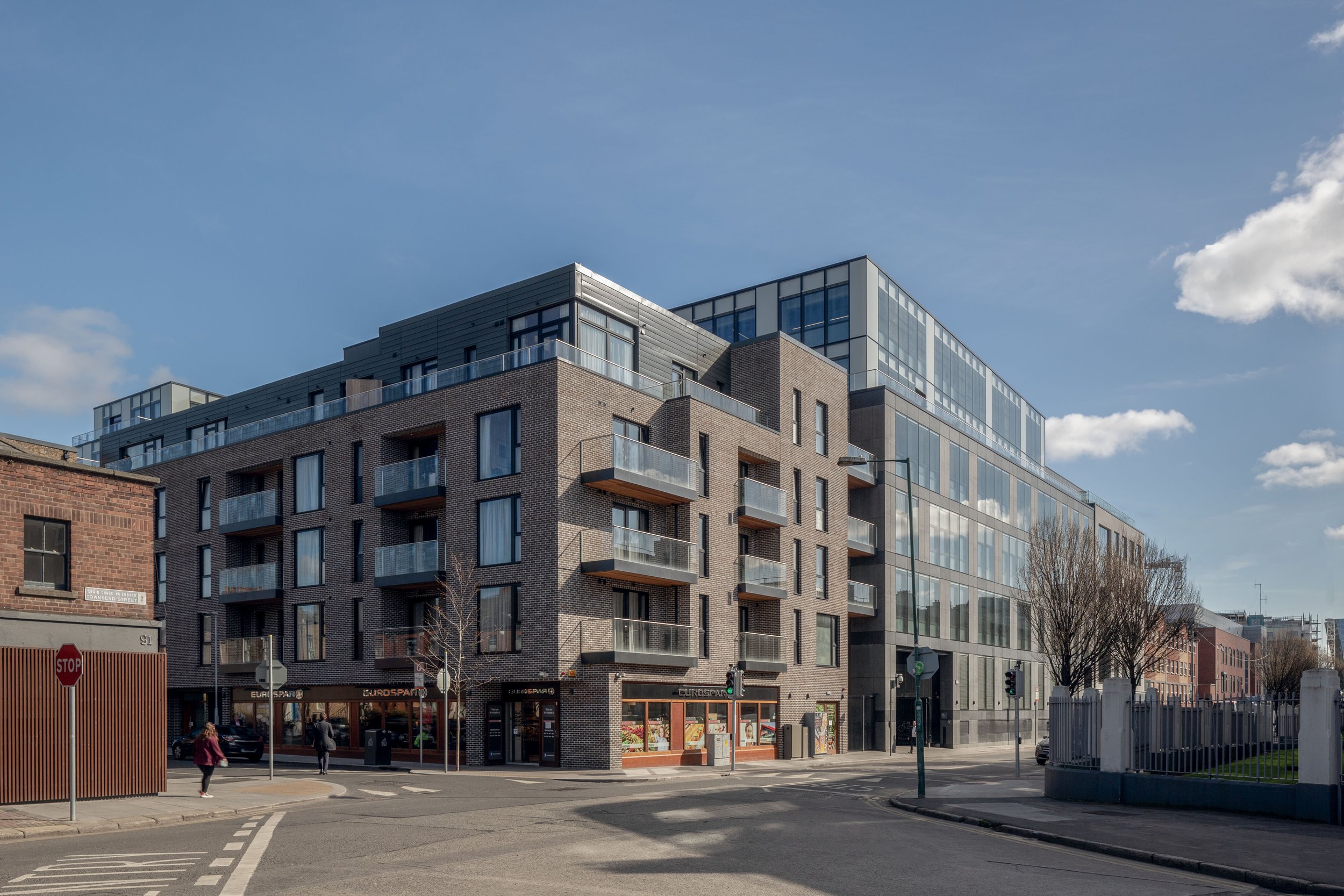 Modern urban building with brick and glass exterior, featuring balconies and ground-level retail stores at an intersection.