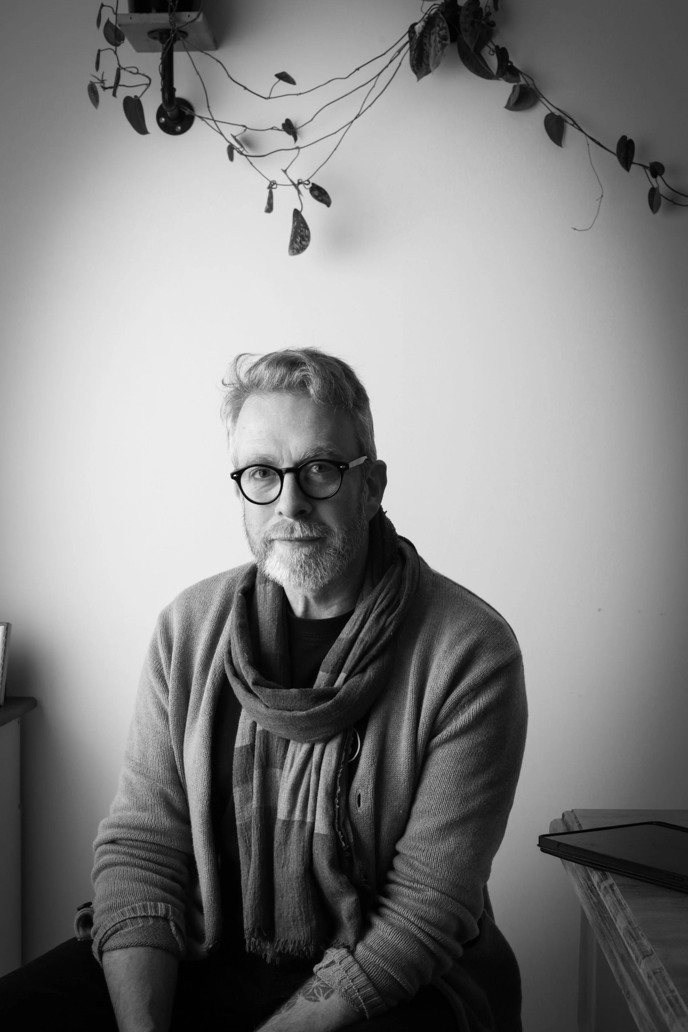A black and white photo of a light-skinned man with glasses, a beard, and short hair, sitting indoors. He's wearing a scarf and a sweater. Above him, there's a potted plant with hanging leaves attached to the ceiling. To his right, there's a desk or table with a closed laptop on it.