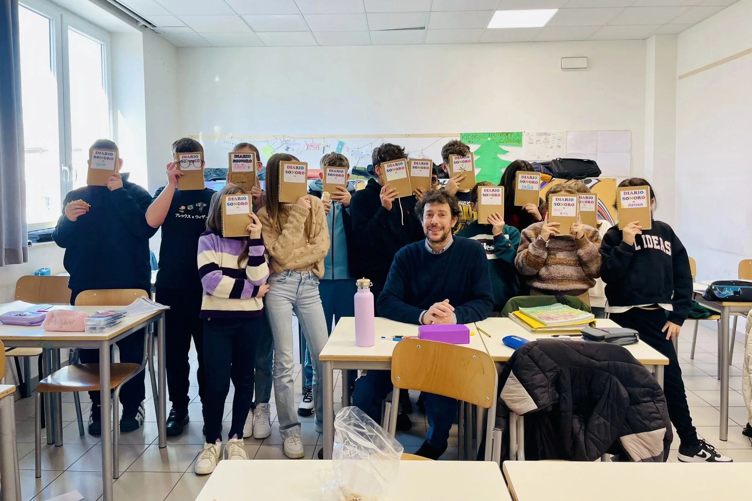Classe di studenti con uno docente, alcuni coprono il volto con quaderni con scritto 'Diario Sonoro', in una classe luminosa con finestra.