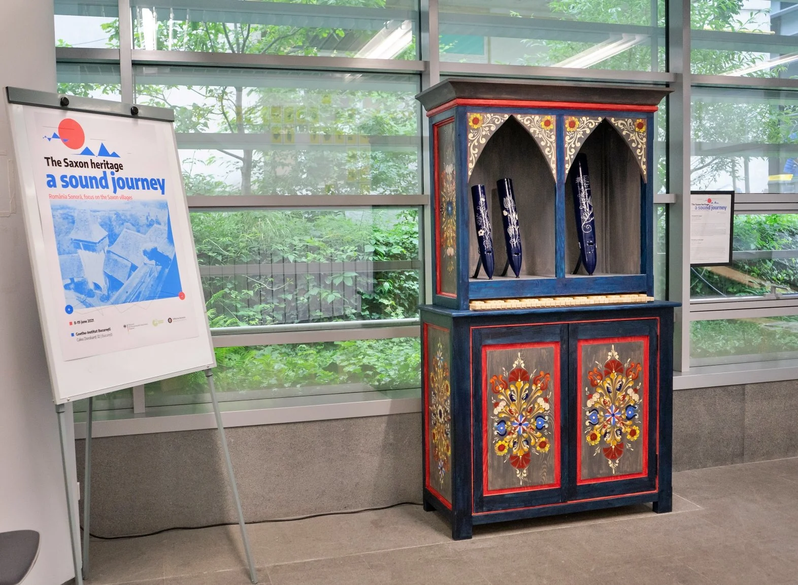 Vetrina decorata con motivi floreali, contenente tre pifferetti blu decorativi. A sinistra, cartellone pubblicitario per un itinerario musicale sulla storia saxon nel sud-est della Romania.