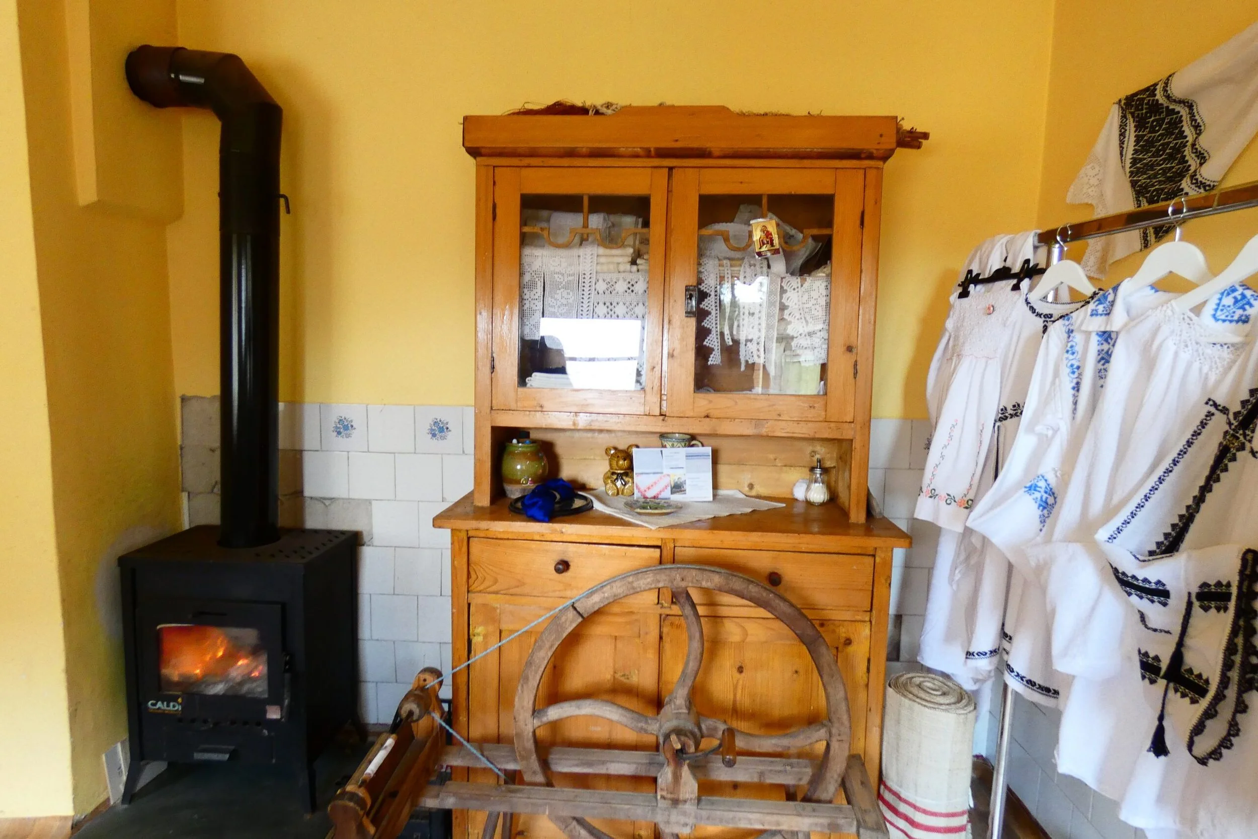 Interno di una stanza tradizionale con armadio in legno, stufa a pellet accesa, e vestiti in stile folkloristico appesi ad un filo.