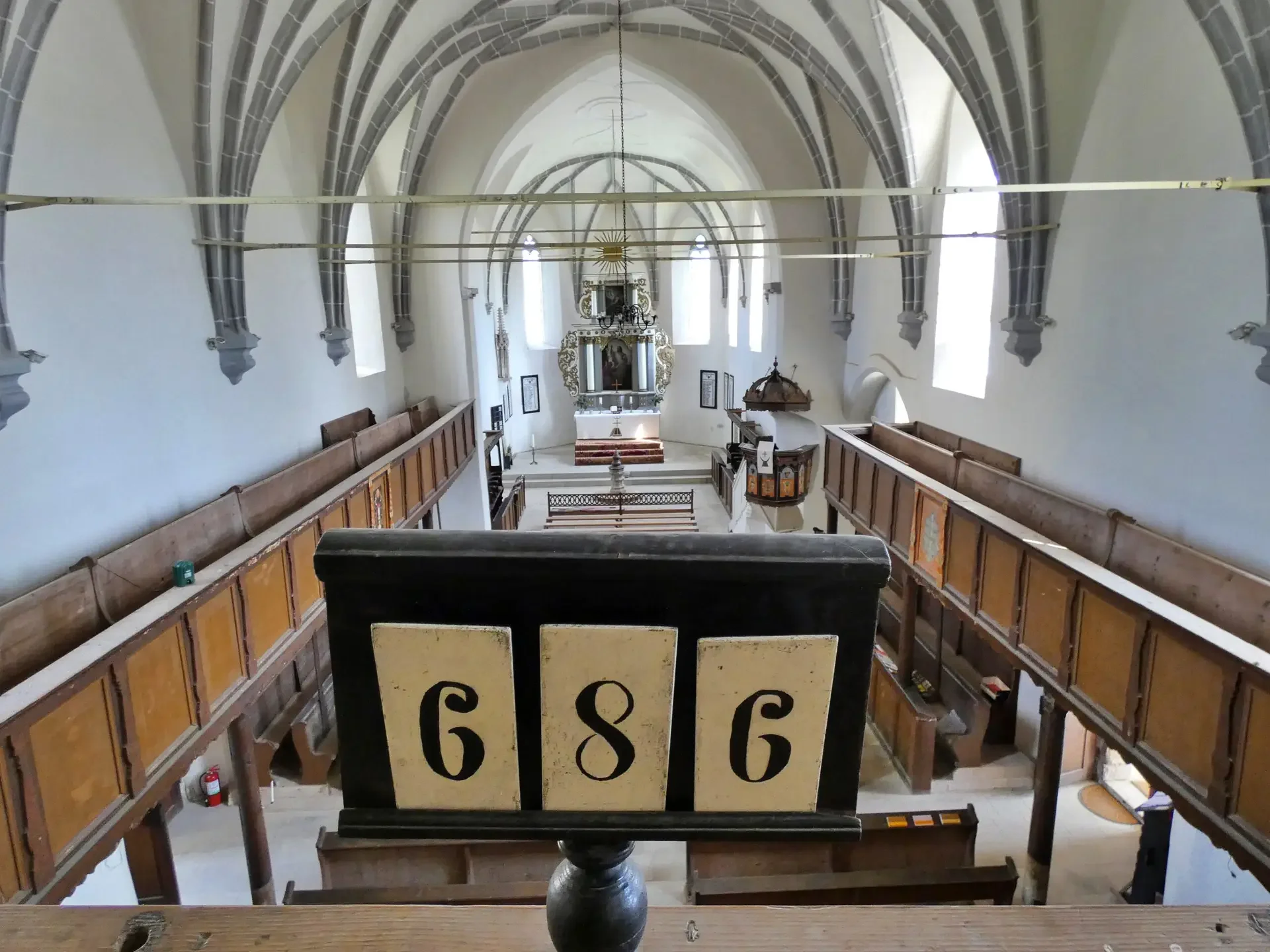 Interno di una chiesa con arcate gotiche, altare decorato e confessional sul lato destro, vista dall'altra balconata con sede vuota e un cartellino con numeri