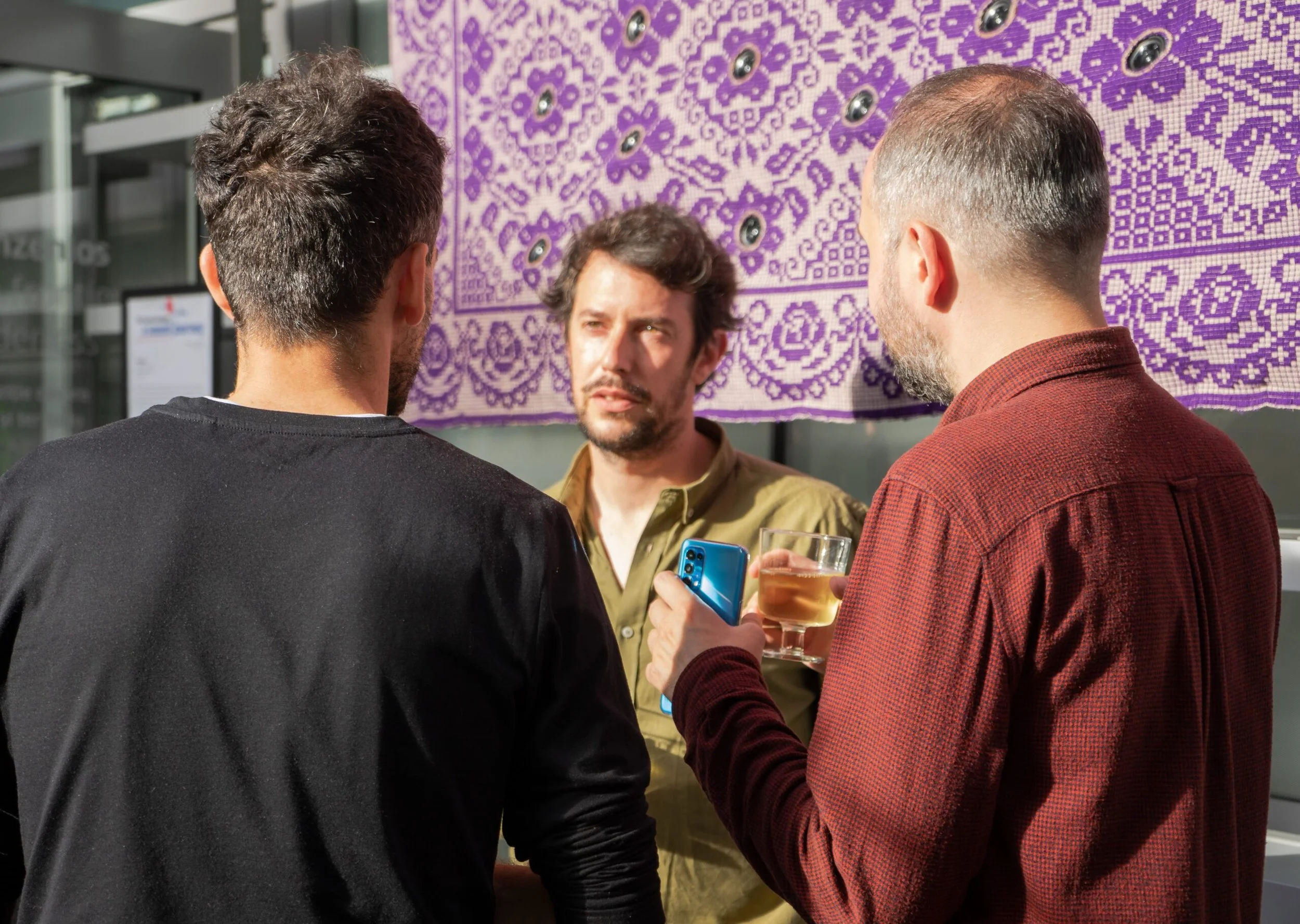 Tre uomini che parlano e ridono, uno dei quali tiene un telefono cellulare blu e un bicchiere di bevanda color ambra, in un'ambientazione urbana con decorazioni violette sullo sfondo.