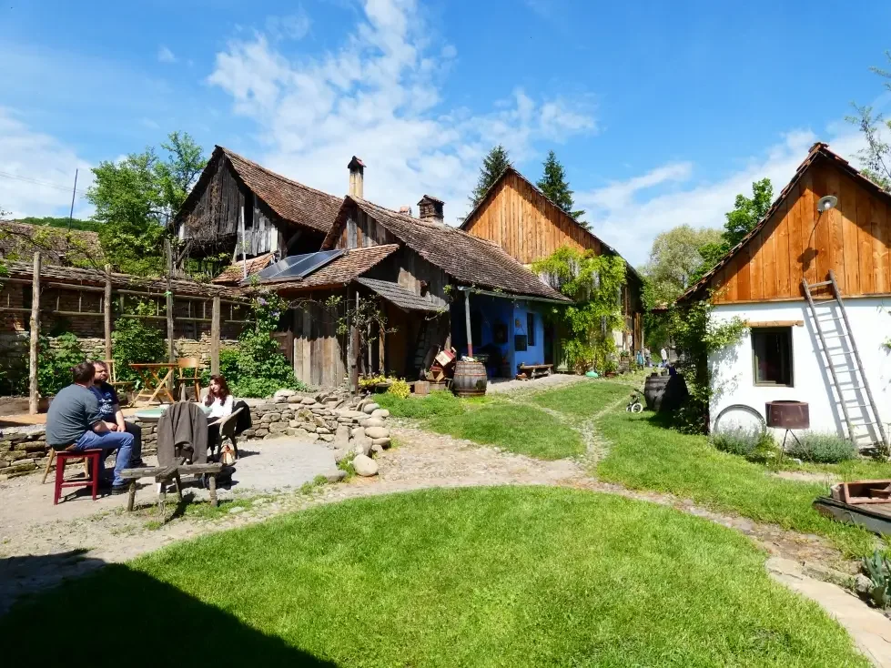Un cortile con alcune persone sedute e alcune case in stile rustico, con tetti di tegole e un cielo azzurro con nuvole.