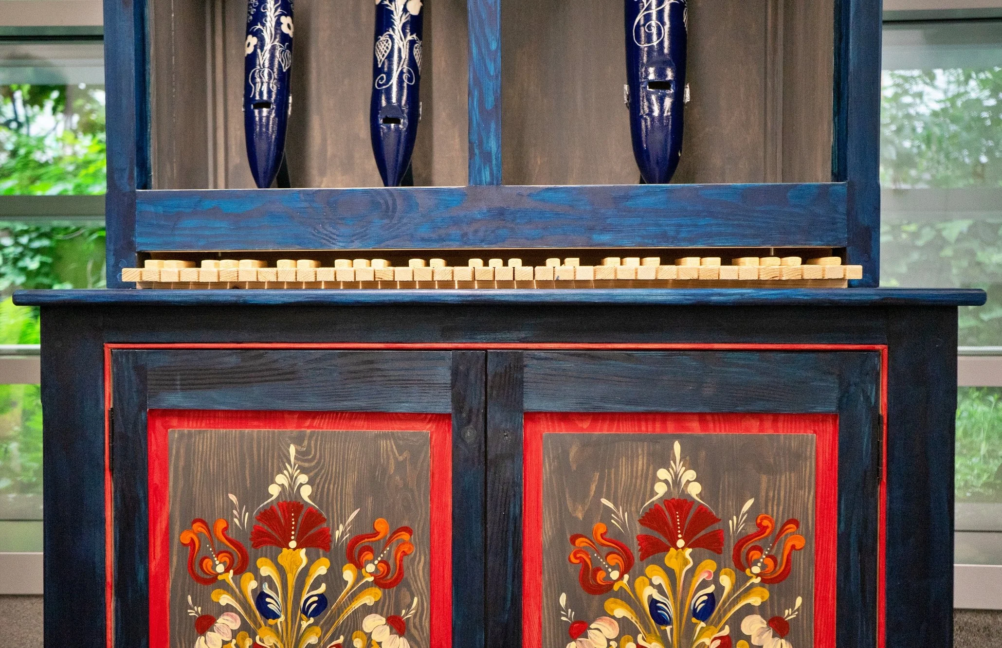 Credenza in legno dipinta con motivi floreali, colori main rosso, blu, giallo e beige, con oggetti in ceramica blu sopra, in una stanza con grande finestra
