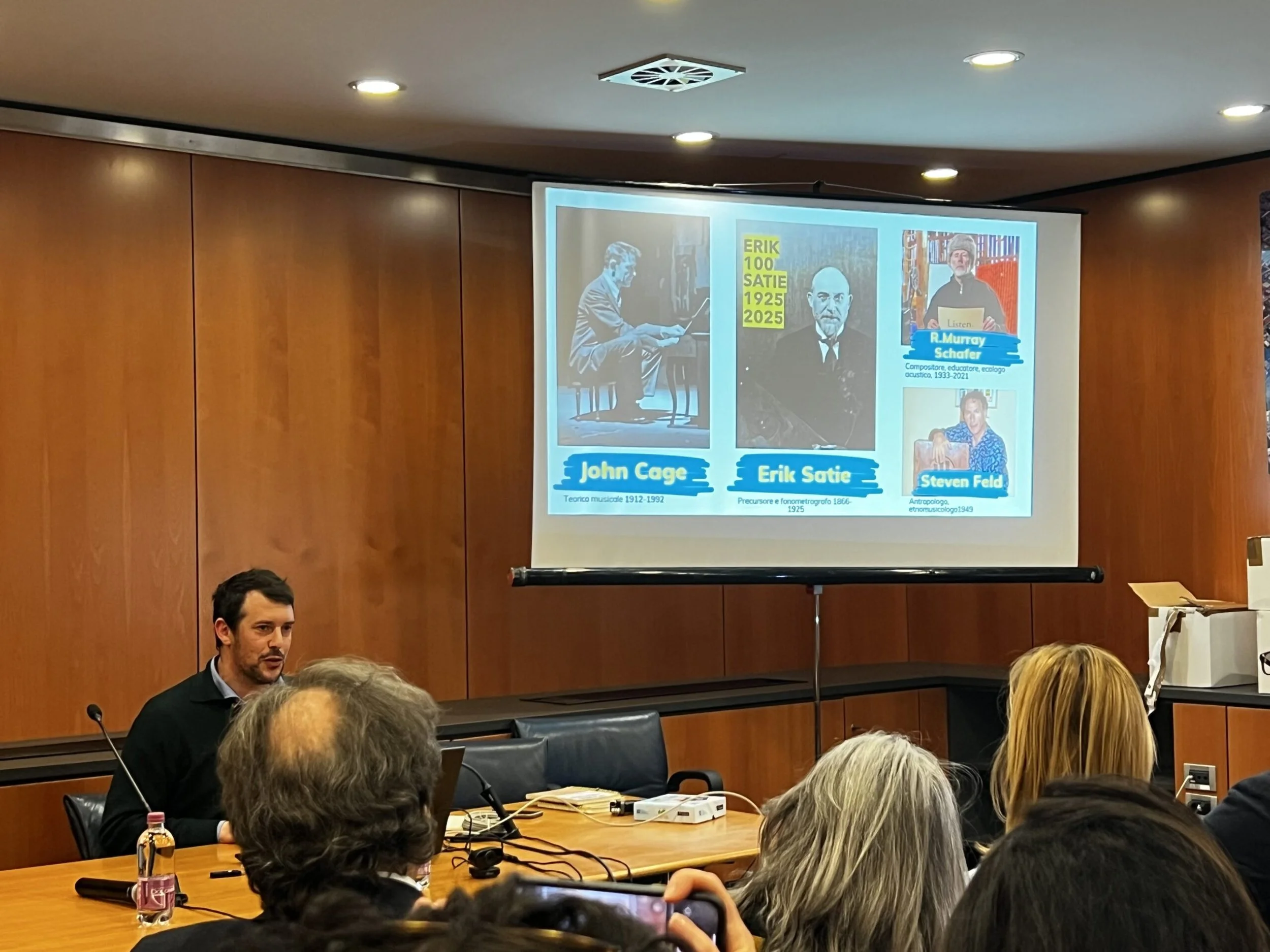 Un uomo seduto davanti a un tavolo durante una presentazione, con pubblico seduto di fronte, proiettore con slide sugli schermi visualizzati dietro di lui.