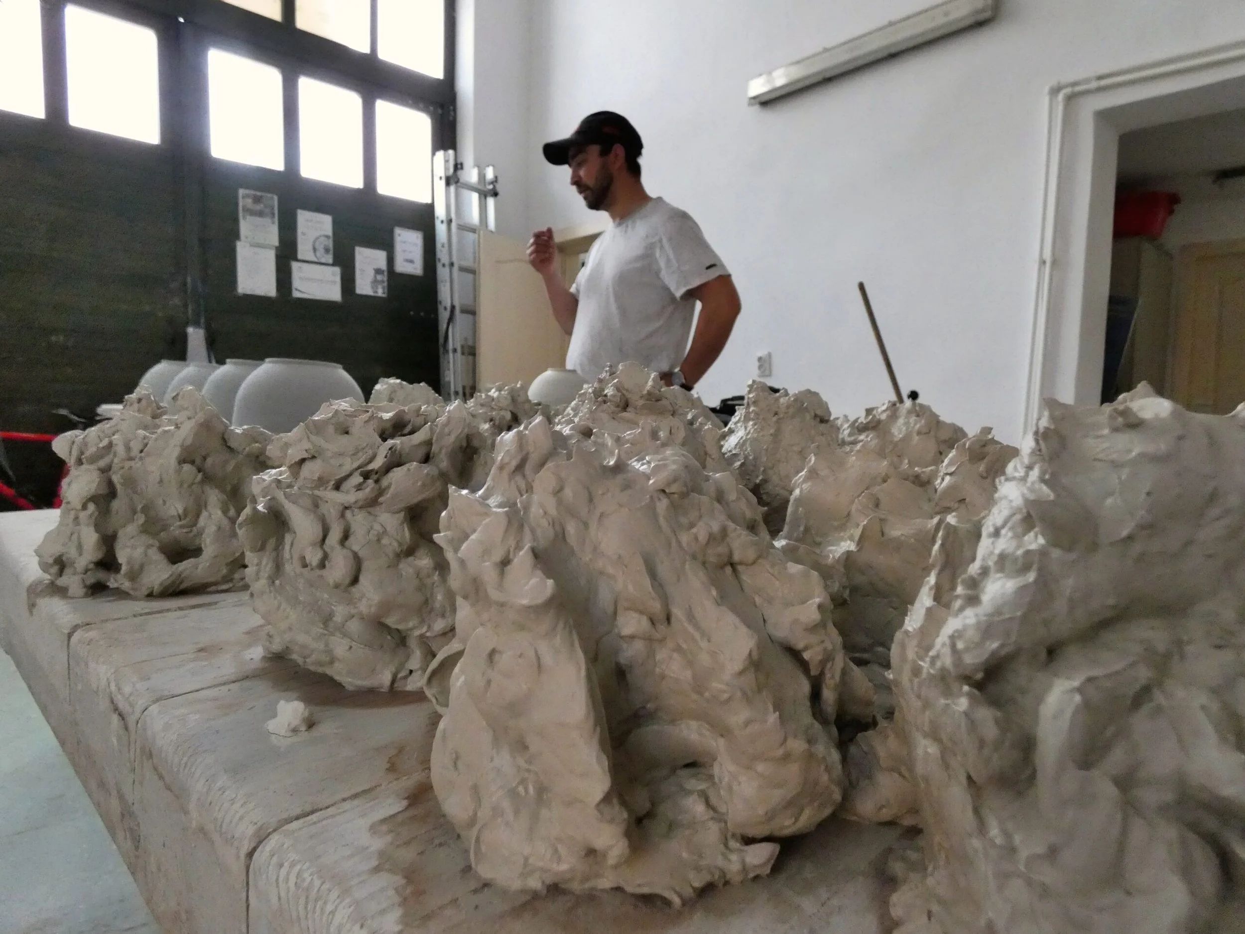 Tavolo con grosse pietre o sculture di argilla, con uomo in piedi dietro che osserva o lavora, all'interno di un ambiente industriale o artistico.