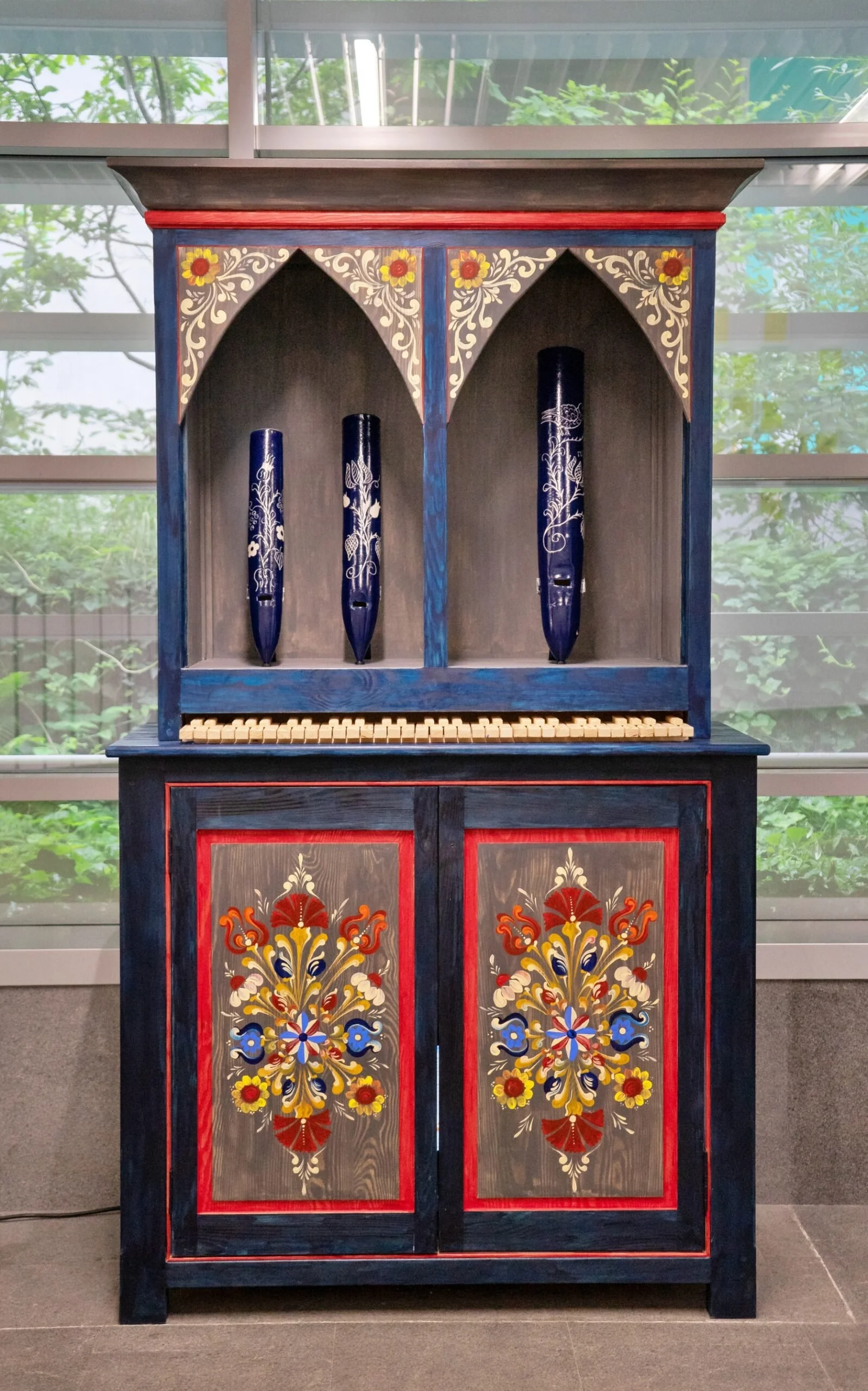 Armadio in legno dipinto con decorazioni floreali colorate, con tre flauti blu decorati, posizionato davanti a una finestra con vista su alberi e foglie.