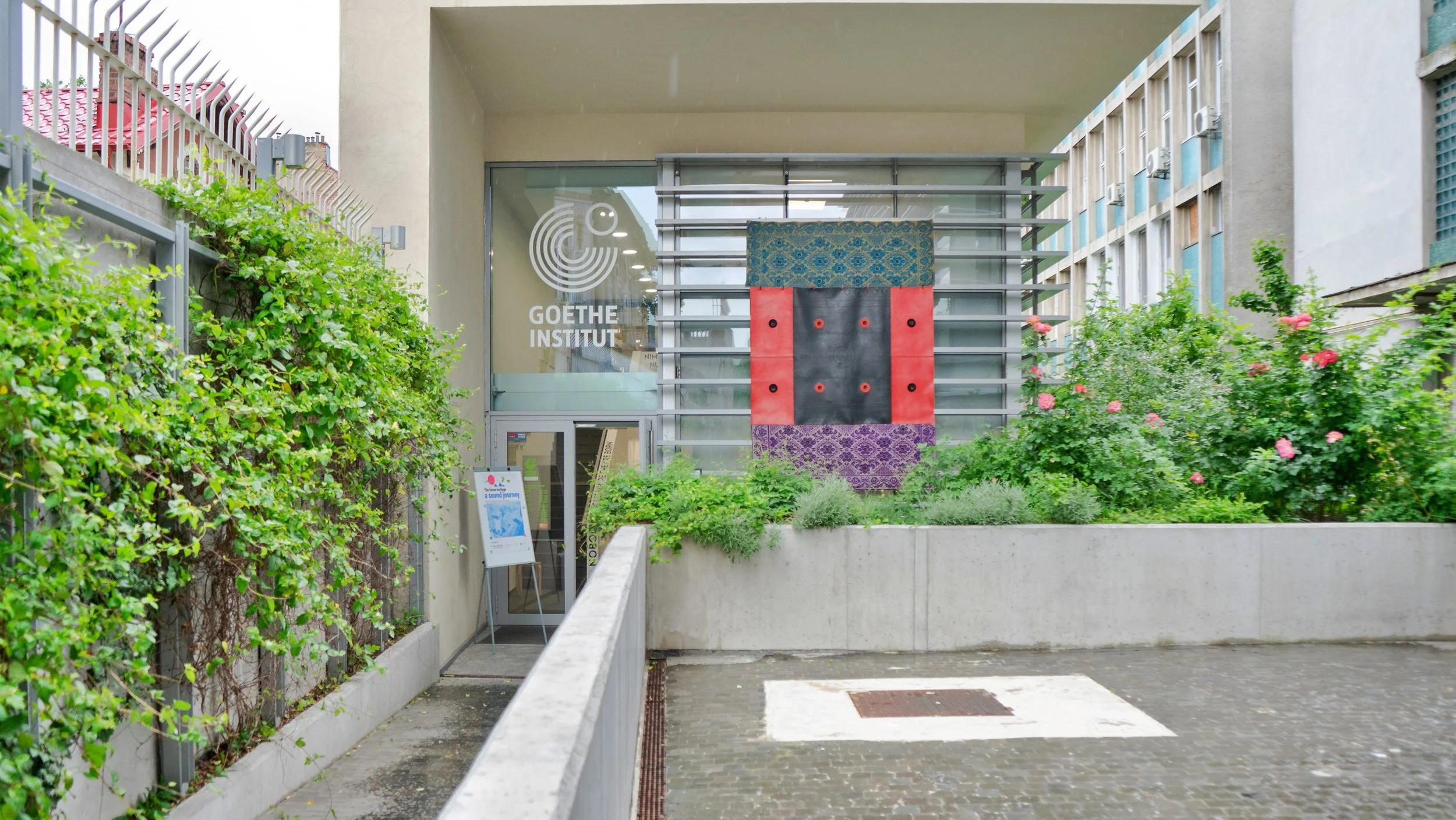 Facciata dell'Istituto Goethe con una vetrata decorata con una grande opera d'arte colorata, circondata da piante e fioriture.