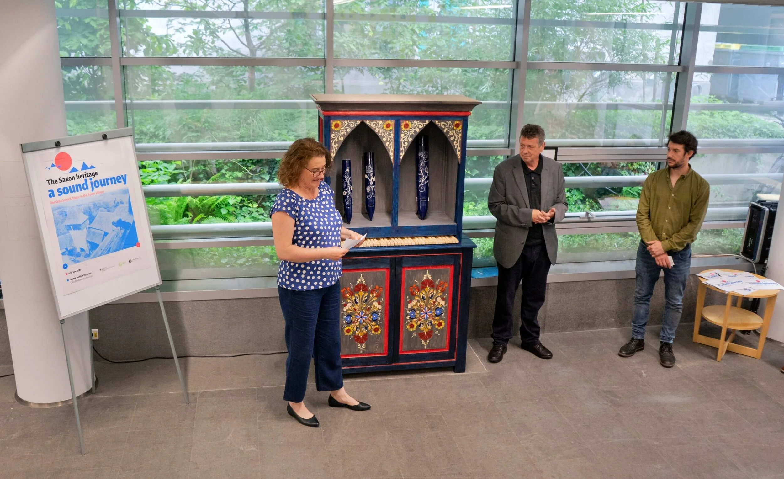 Tre persone in una stanza con grandi finestre di vetro che mostrano una vegetazione verde fuori, una donna che legge da un foglio, e due uomini che ascoltano attentamente, con una credenza decorata tra loro e un cartellone con scritto 'a sound journe