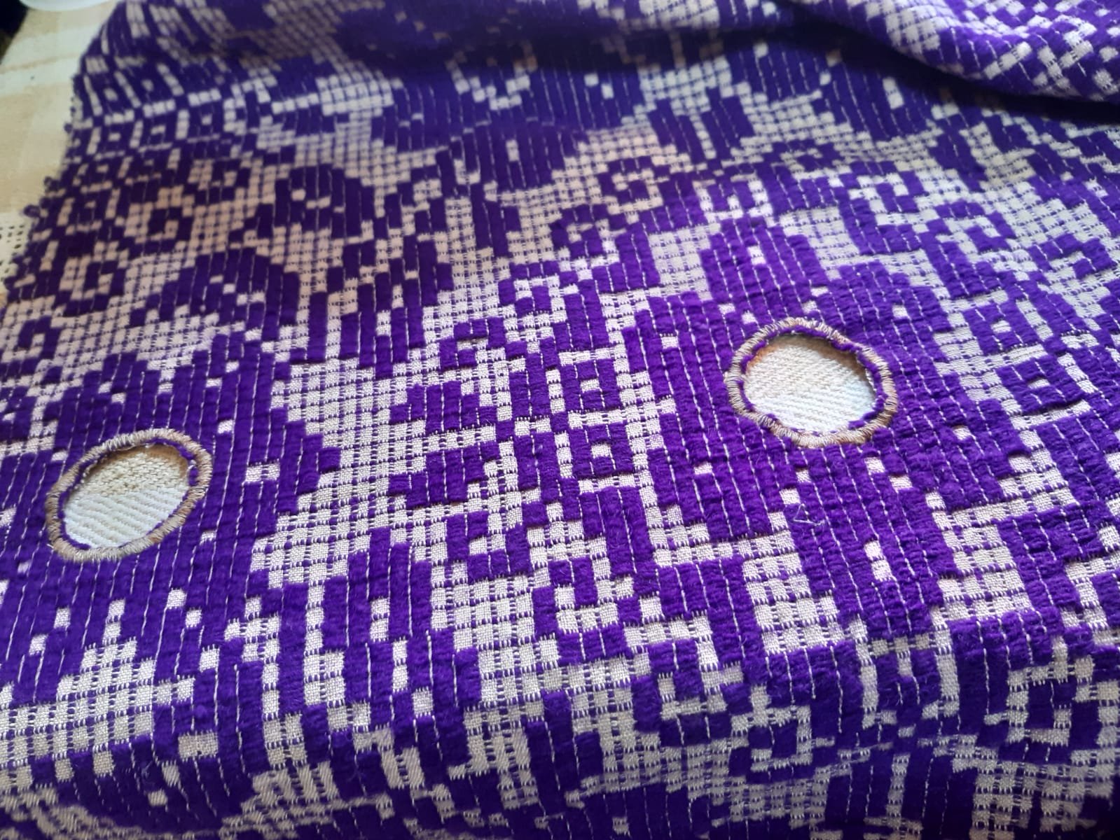 Una stoffa ricamata con dettagli viola e bianchi, con due fori rotondi e un motivo geometrico complesso.