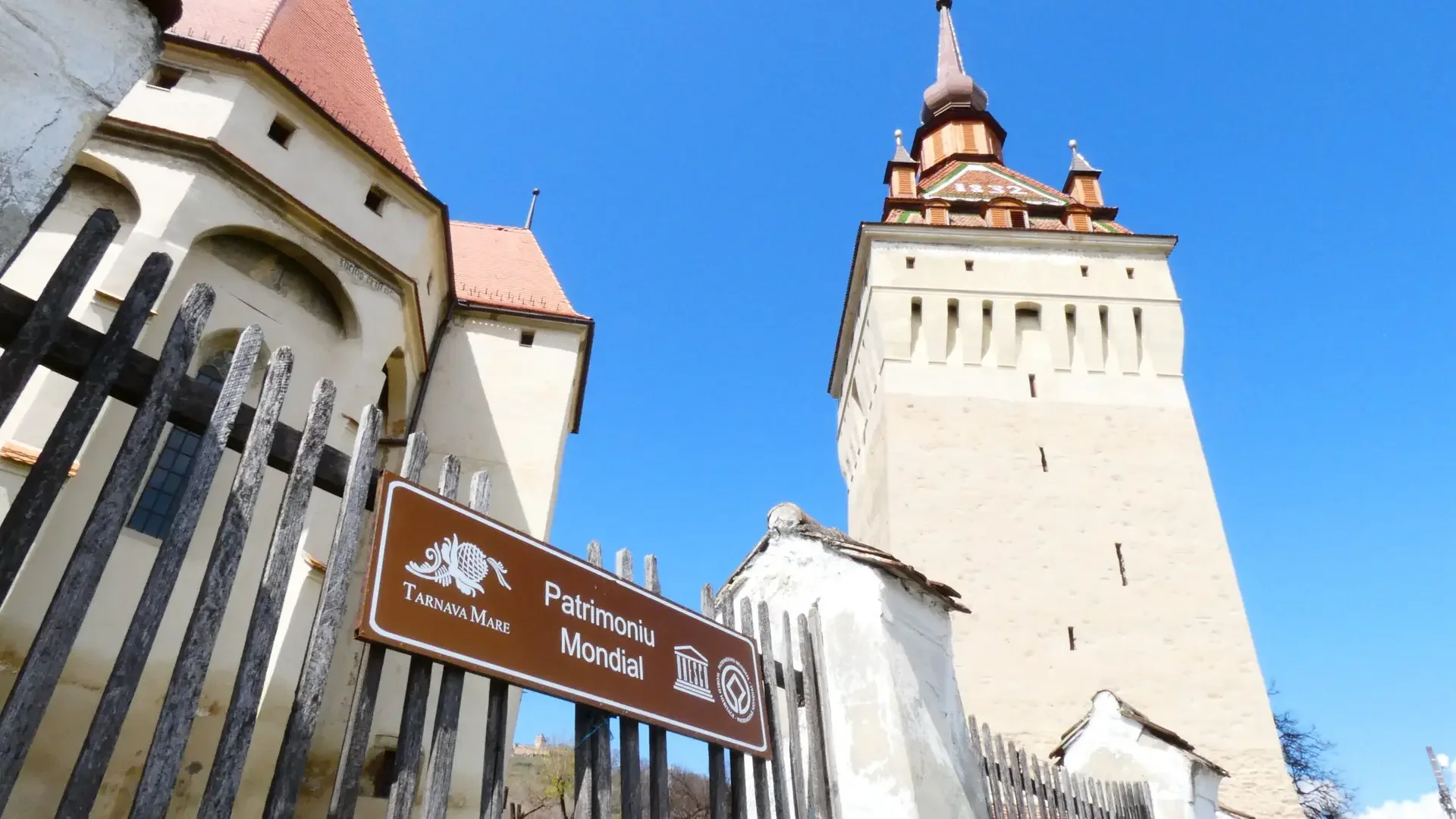 Immagine di un castello con torri, con un cartello marrone con scritte in italiano e simboli di patrimonio mondiale, recante il testo 'Patrimonio Mondial' e 'Tarnava Mare', e un cancello in ferro arrugginito davanti al castello, sotto un cielo azzurr