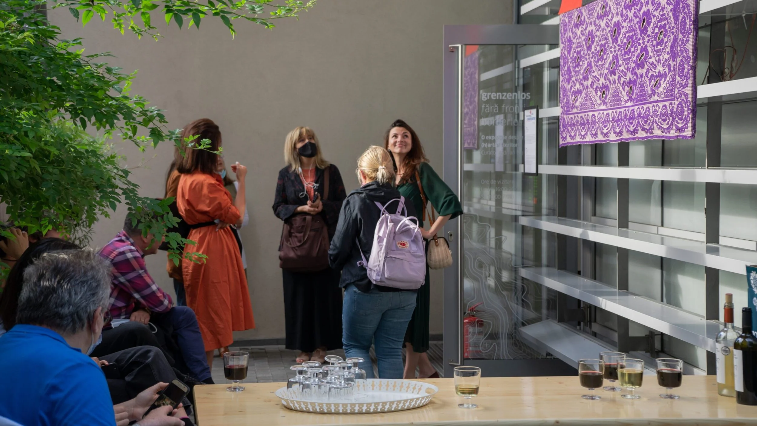 Gruppo di persone in attesa di entrare in una mostra, alcune sedute e altre in piedi, tra cui una donna con una borsa beige che parla con altre donne. Sul tavolo ci sono bicchieri con vino e acqua, e sulla parete di sfondo è appeso un tessuto viola c