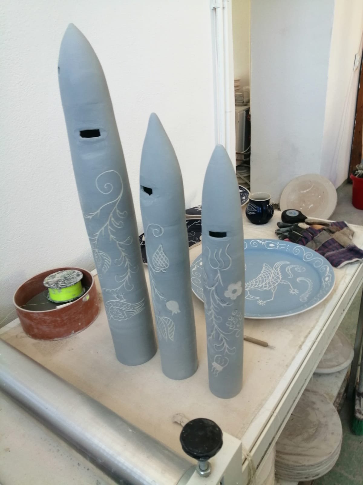 Tre astucci di forma allungata e di colore grigio con decorazioni floreali, posizionati in verticale su una superficie di lavoro, oltre a vari strumenti e oggetti di artigianato in un'area di lavoro.