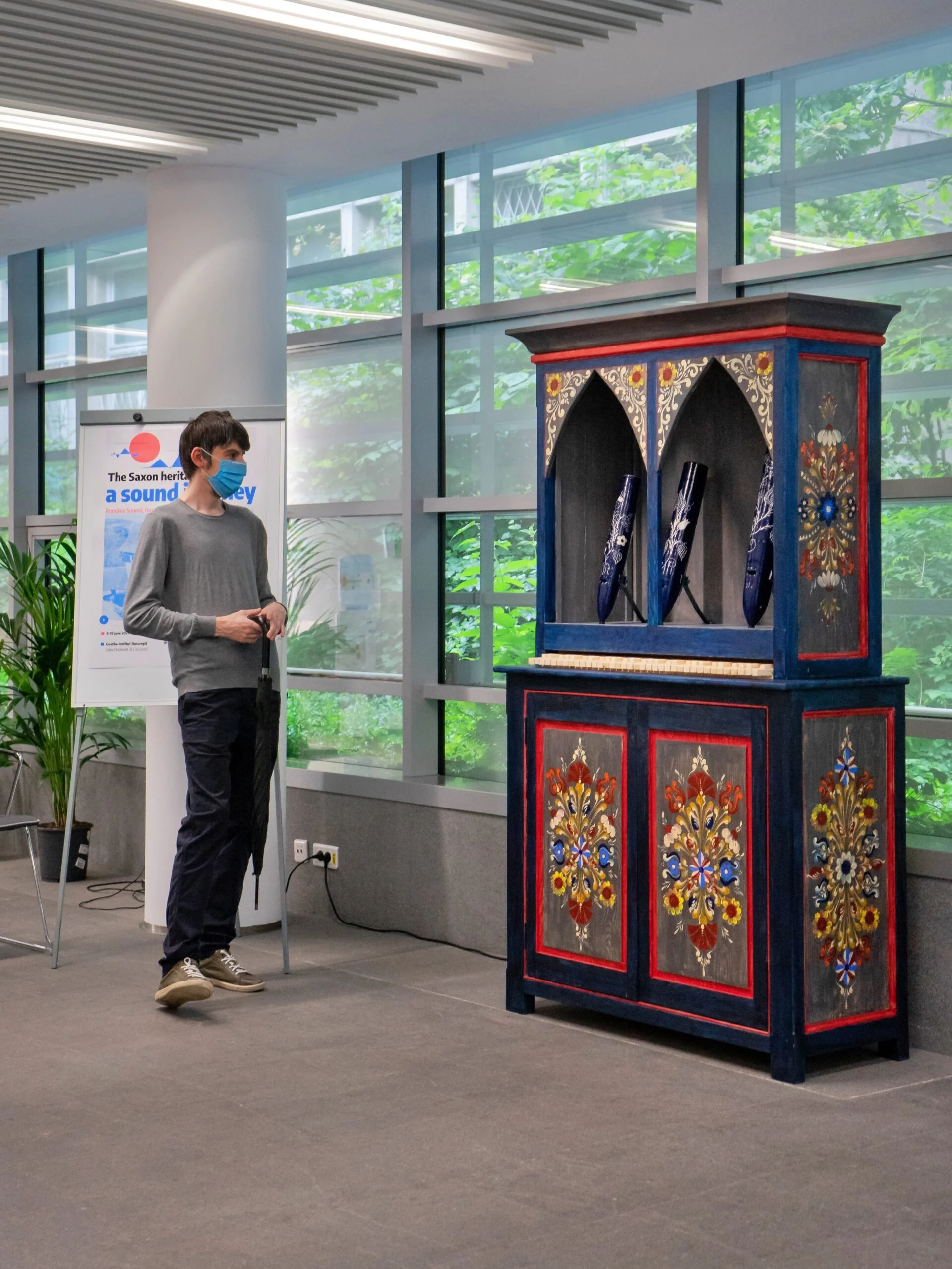 Un uomo con una mascherina che tiene un ombrello, osserva uno strano organo musical raffigurato come un mobile dipinto con motivi floreali colorati, in un ambiente con grandi finestre e piante.