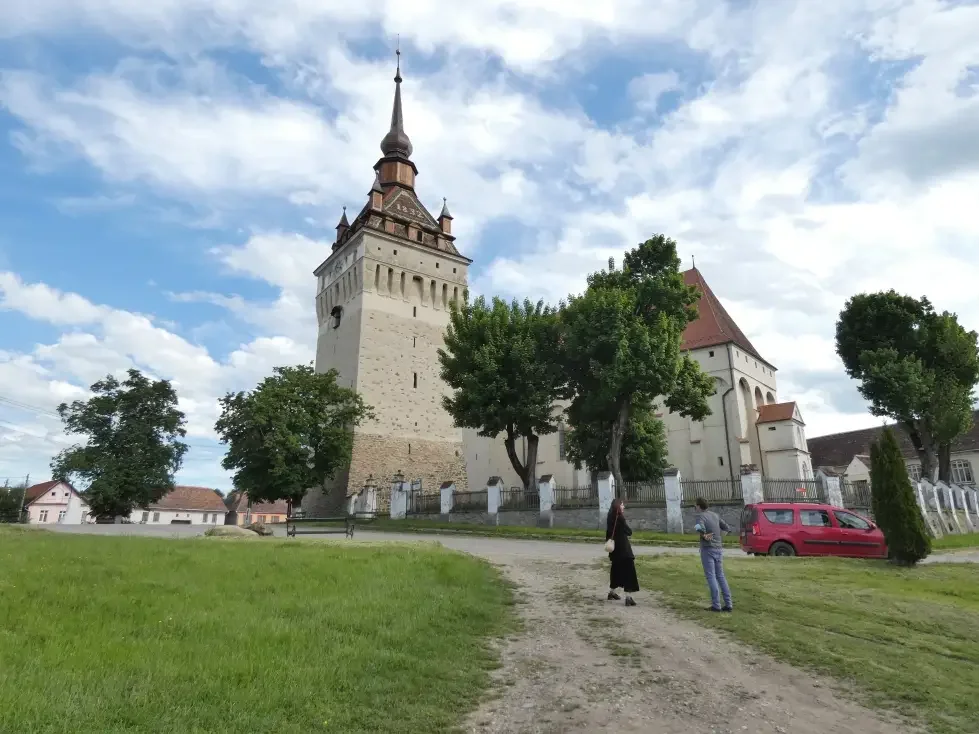 Una chiesa storica con torre alta, circondata da alberi e un muro di cinta, con un'area erbosa davanti e due persone che parlano vicino a una vettura rossa.