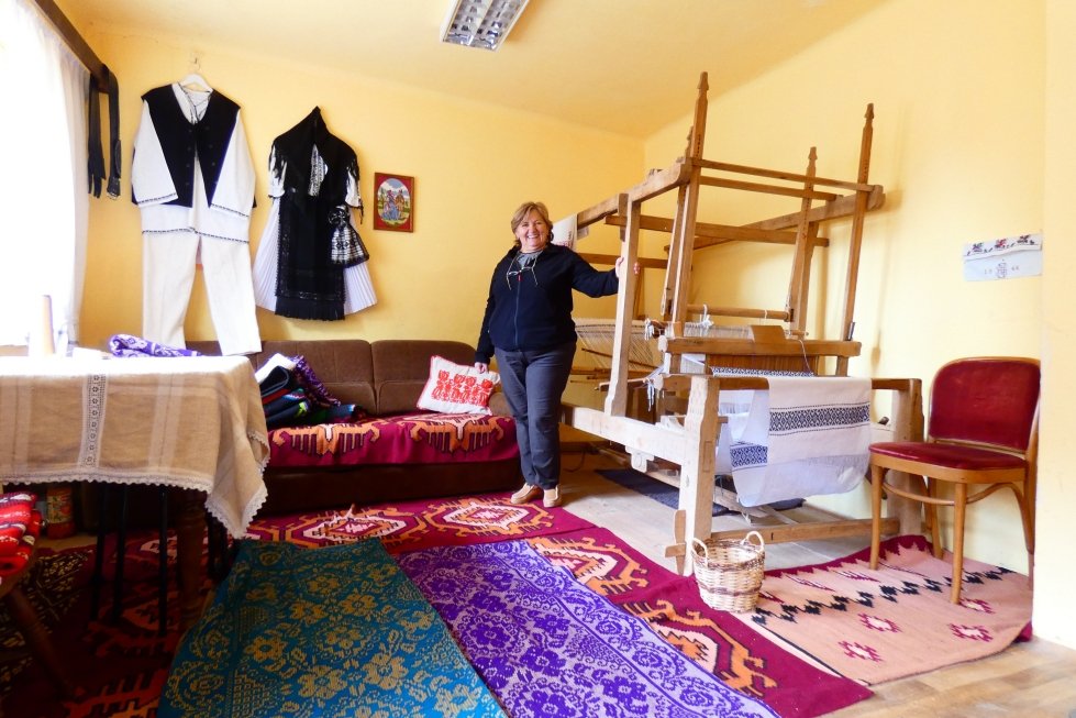 Una donna sorridente in una stanza che mostra un telaio per tessere a sinistra e arredi tradizionali, con tappeti colorati e vestiti appesi al muro.
