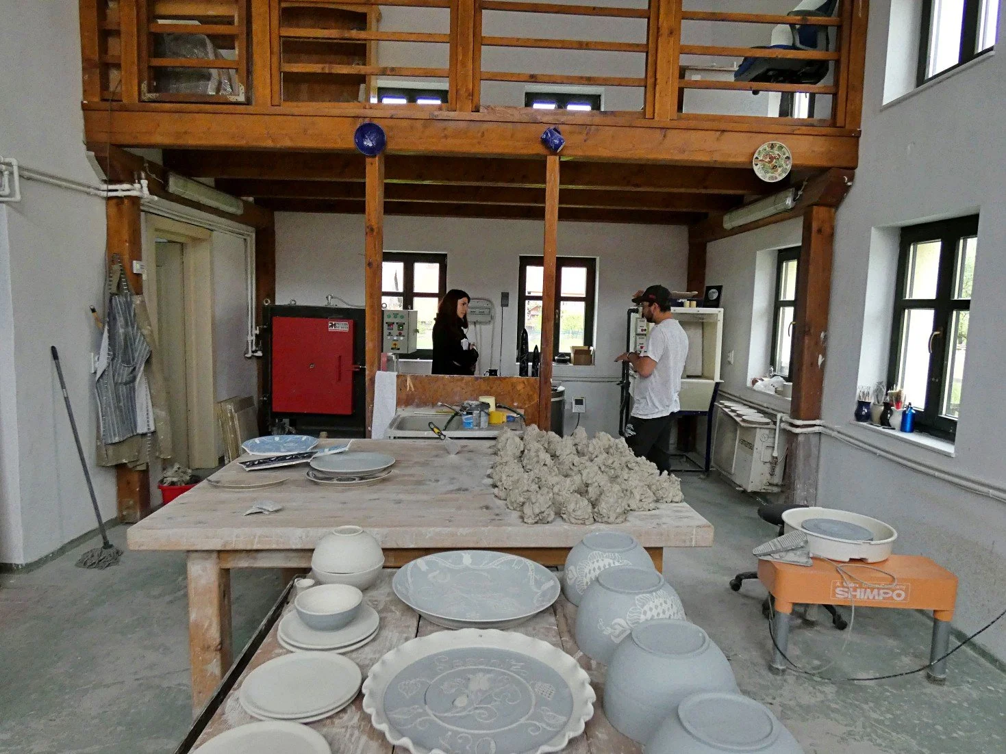Interno di un laboratorio di ceramica con tavoli di lavoro, ceramiche in varie fasi di lavorazione, due persone che parlano al centro, scaffali e utensili, balcone con finestre