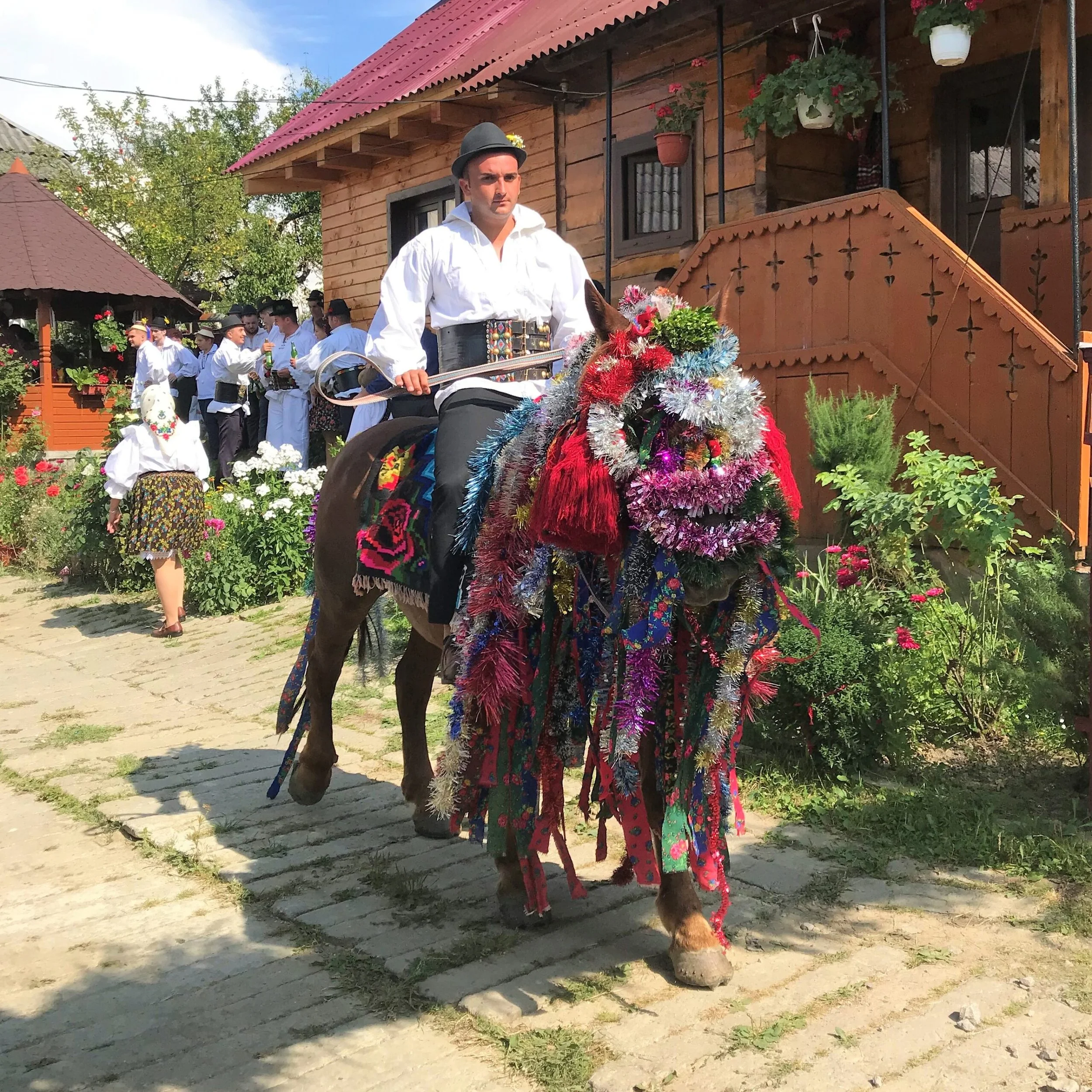 Uomo vestito con abiti tradizionali su un cavallo decorato con fiori e nastri colorati, in un cortile con altri partecipanti a una festa tradizionale.