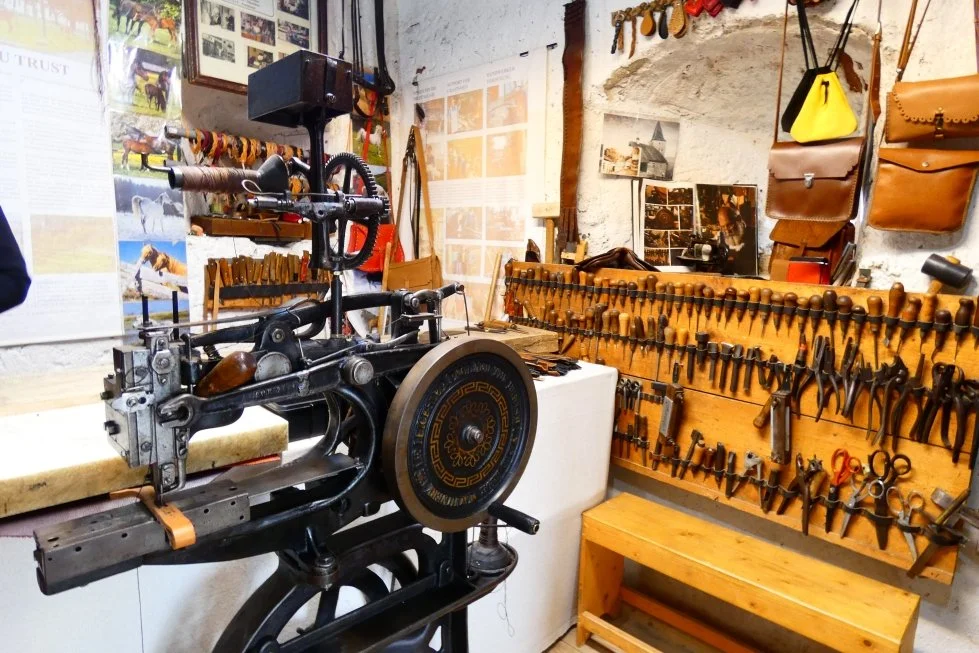 Un laboratorio artigianale con attrezzi per la lavorazione del cuoio, tra cui martelli, scalpelli e strumenti di pinzatura appesi alle pareti e su un pannello di legno, con una macchina da cucire in primo piano.