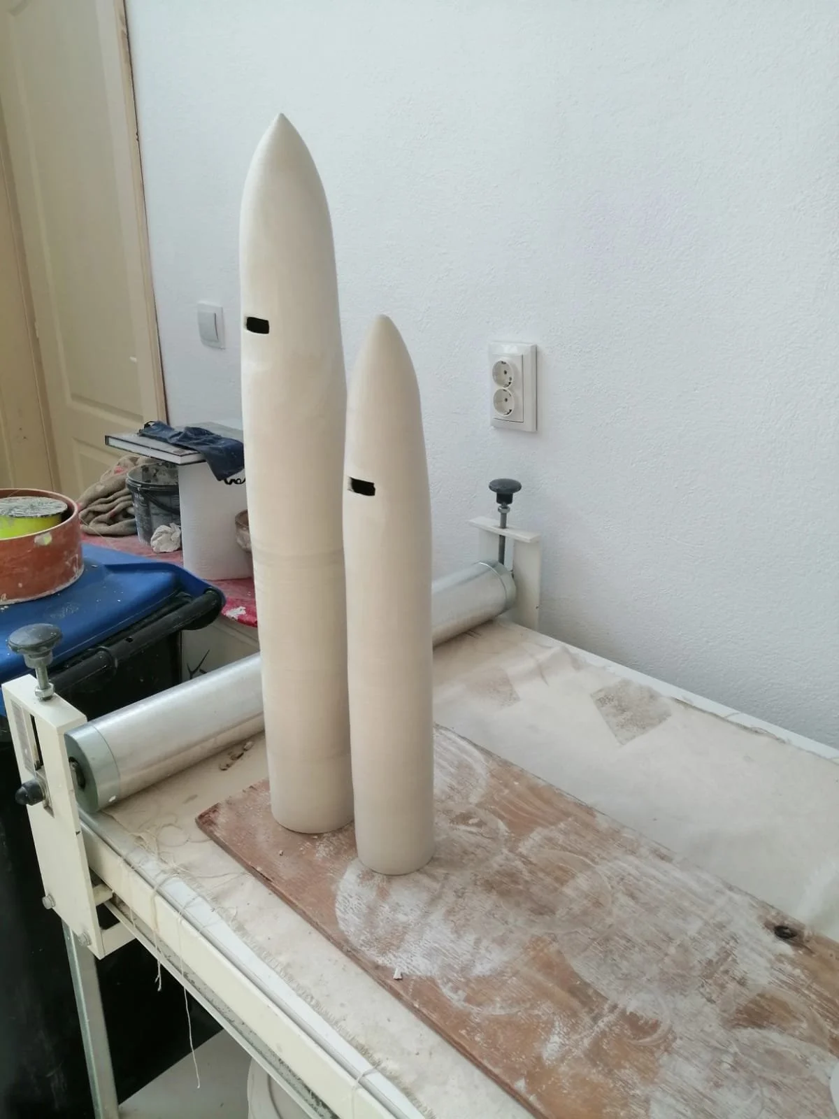 Due modelli di missili di falegnameria o di plastilina, uno più grande e uno più piccolo, in fase di modellatura su un banco di lavoro, con strumenti e materiali intorno.