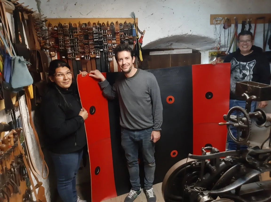 Tre persone in un laboratorio di strumenti musicali, con strumenti appesi alle pareti e tre pannelli colorati davanti a loro. La scena è accogliente e informale.