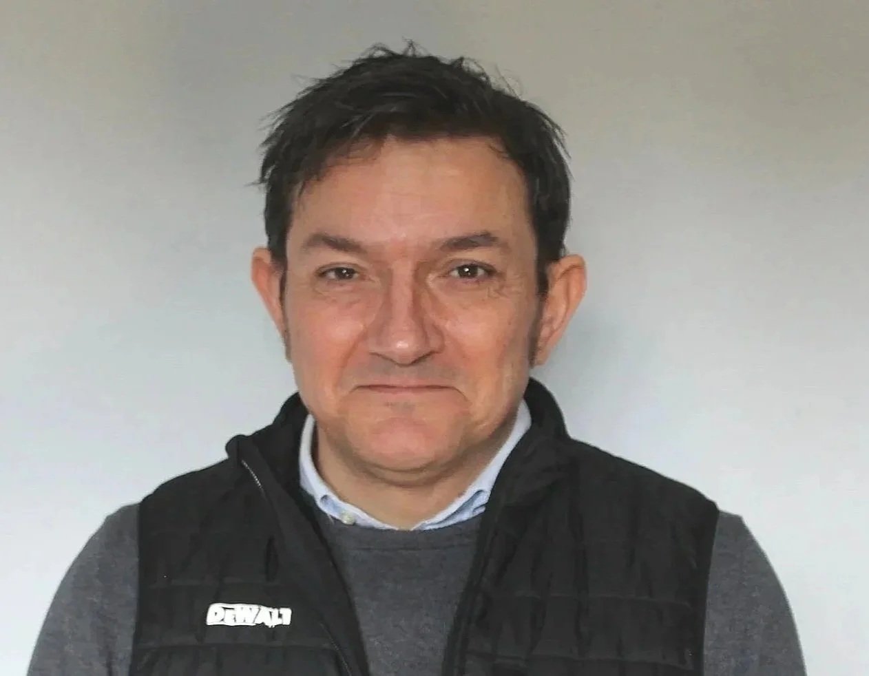 A middle-aged man with dark hair and light skin wearing a dark jacket over a gray sweater and collared shirt, standing against a plain wall.