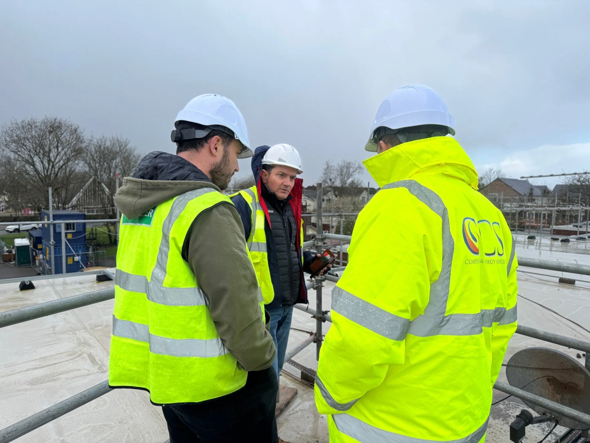Three construction workers wearing high-visibility jackets and hard hats standing on a rooftop, having a discussion, with cloudy sky and residential houses in the background.
