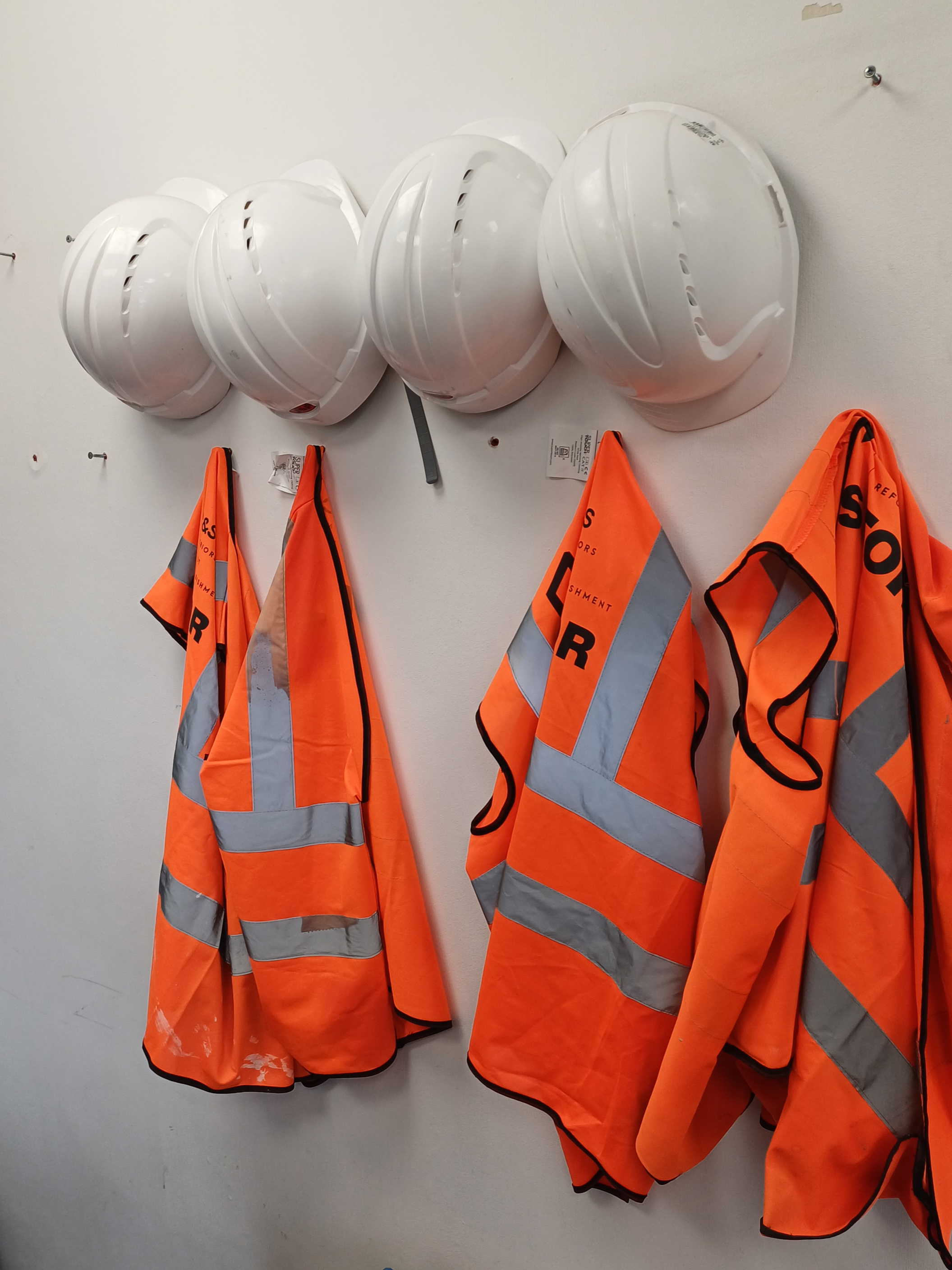 Five white hard hats mounted on a wall above orange safety vests with reflective stripes.