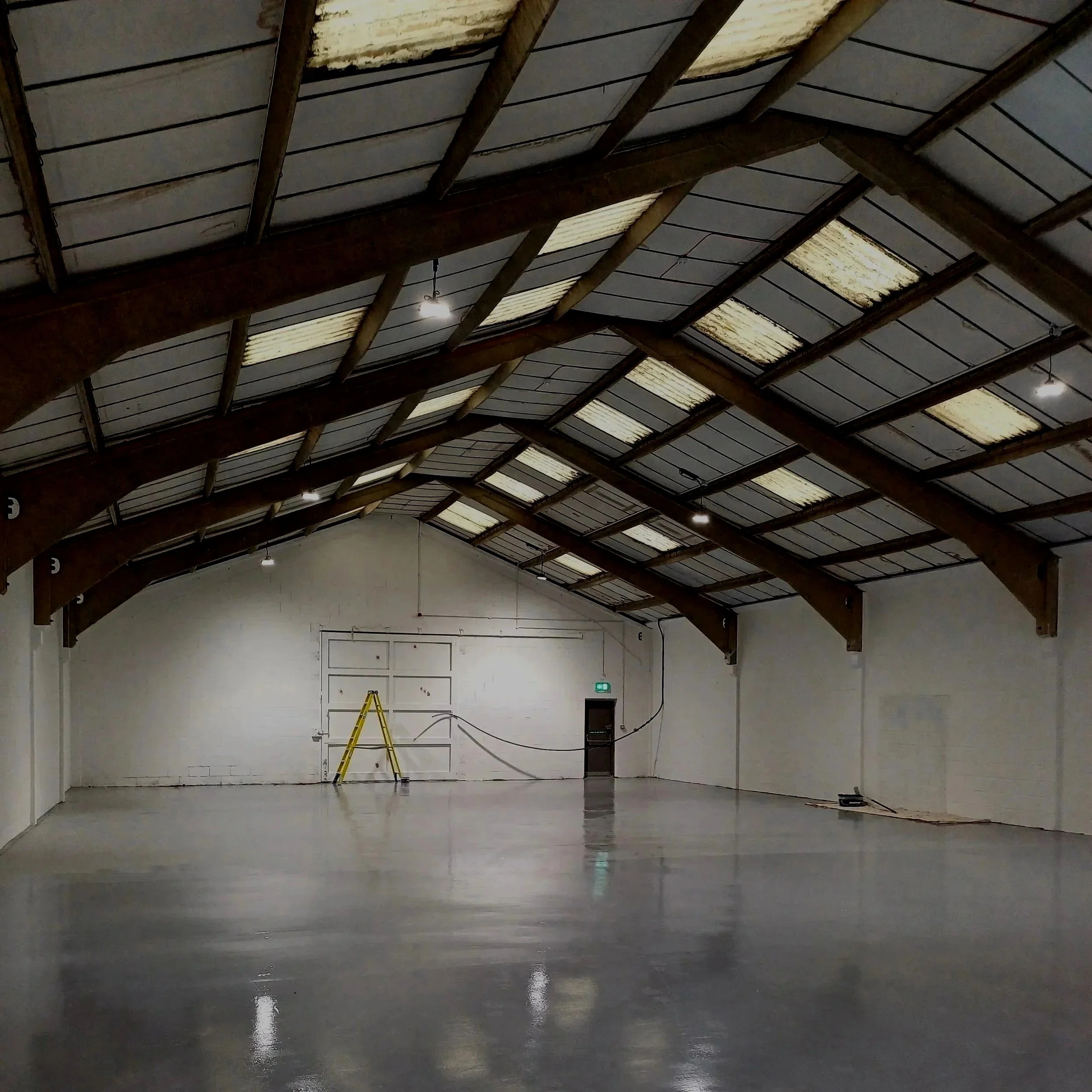 An empty large indoor space with a polished concrete floor, high ceiling with exposed wooden beams, and skylights letting in natural light. There is a yellow ladder and some small electrical cords on the floor near the back wall, which has a ladder and a green exit sign.