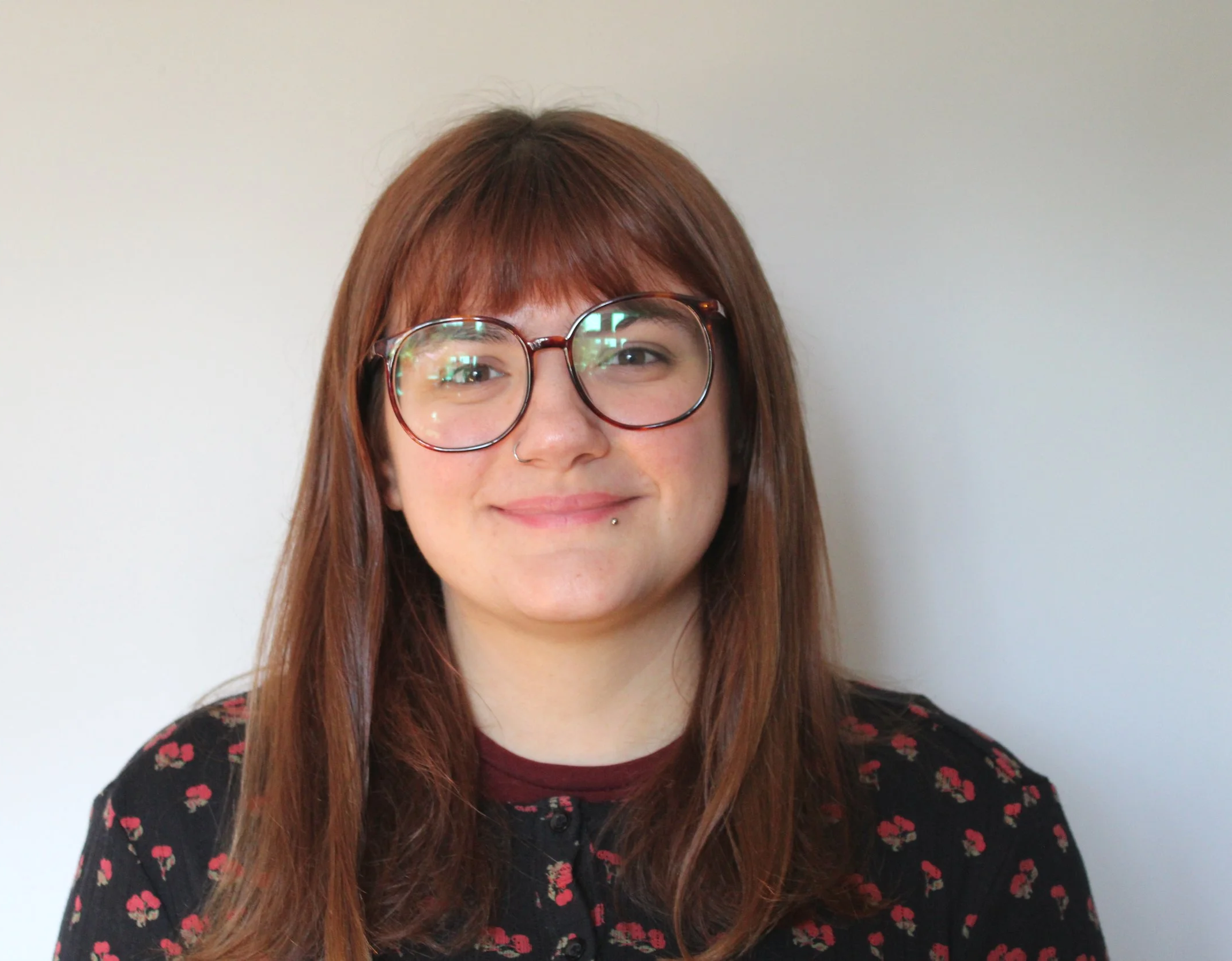 A woman with long reddish-brown hair, round glasses, and a nose piercing smiling in front of a plain light-colored wall.
