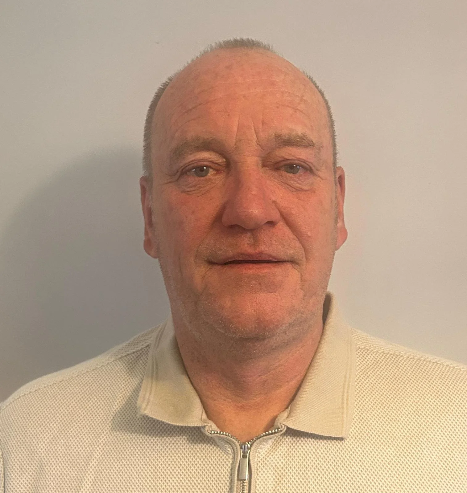 Close-up of a middle-aged man with short, light gray hair, wearing a beige collared shirt with a zipper. He is standing in front of a plain, light-colored wall.