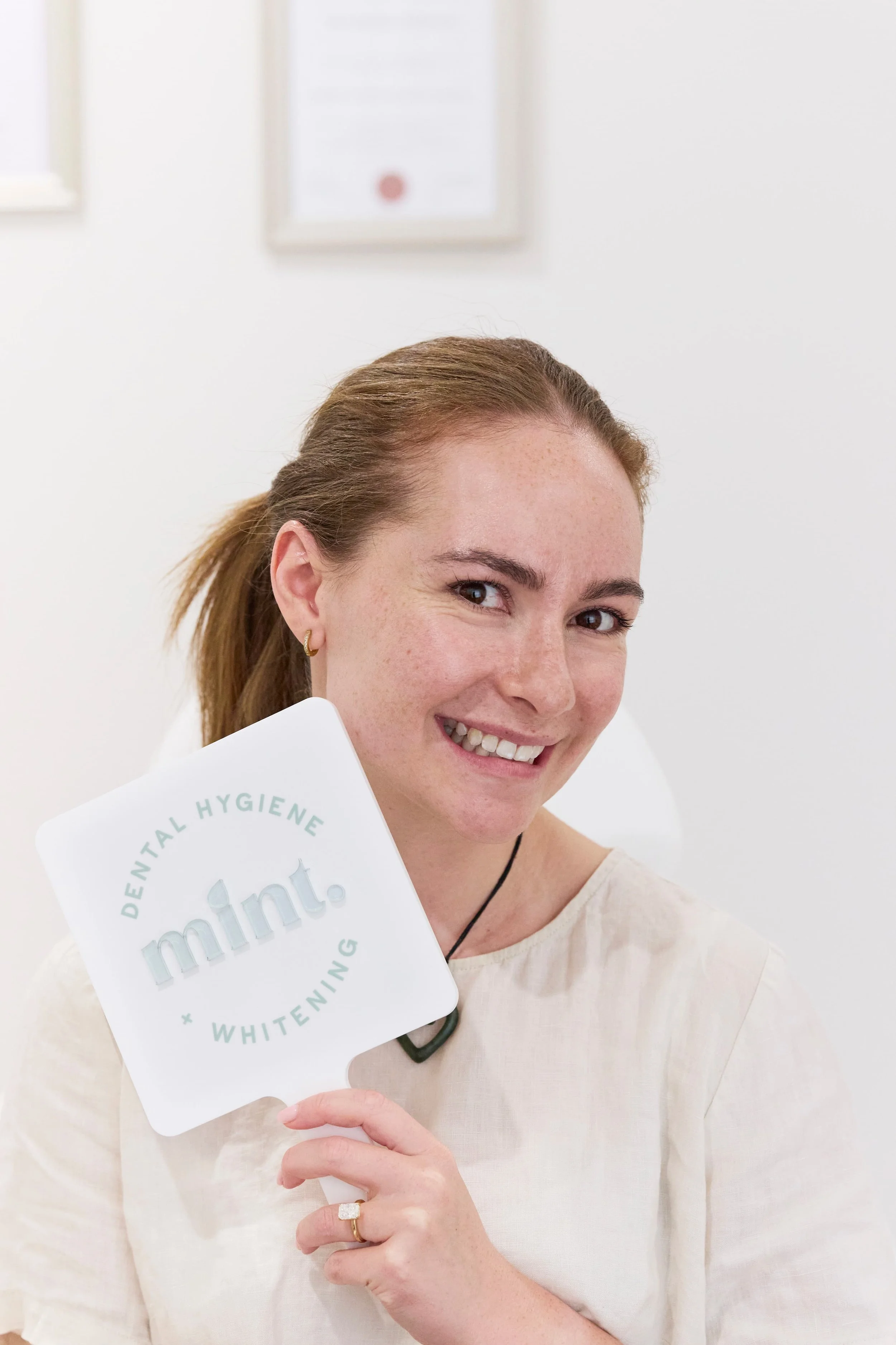A smiling woman with reddish-brown hair and earrings holding a white sign that says 'dental hygiene, whitening + whitening' in a bright, minimal office space with framed documents on the wall behind her.