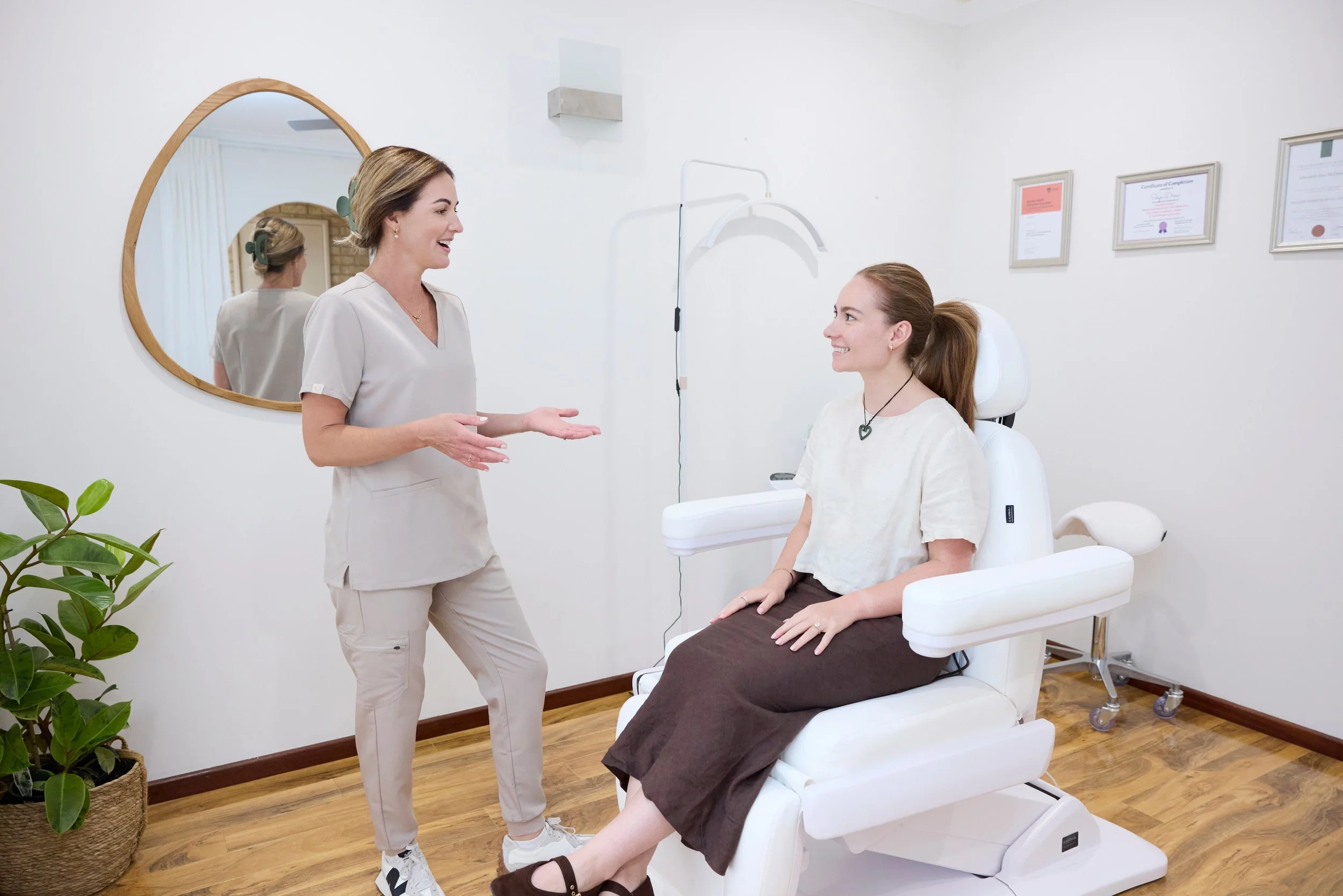 A healthcare professional talking to a woman sitting in a medical examination chair in a clinic room with framed certificates on the wall