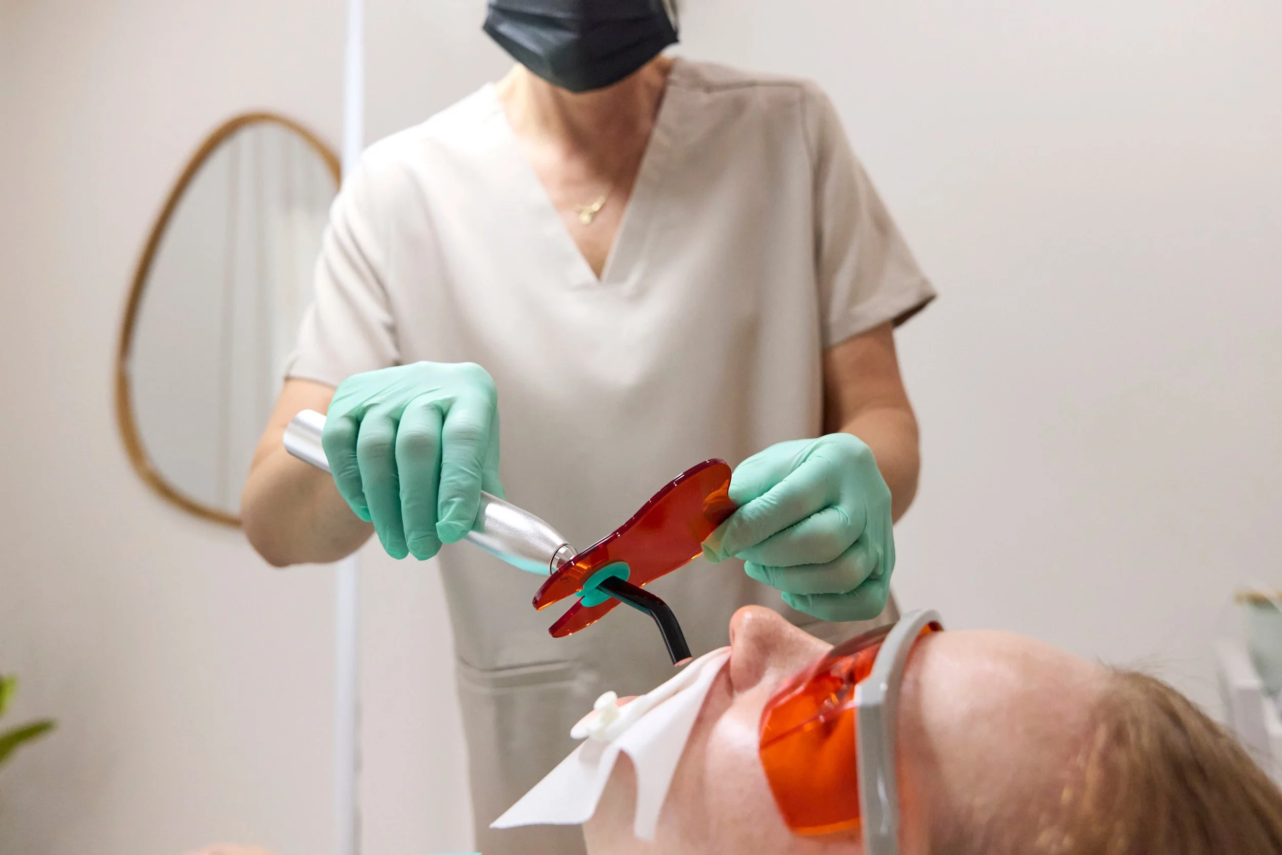 A professional dental hygienist wearing a black mask, beige scrubs, and green gloves holding a medical syringe and a vial.
