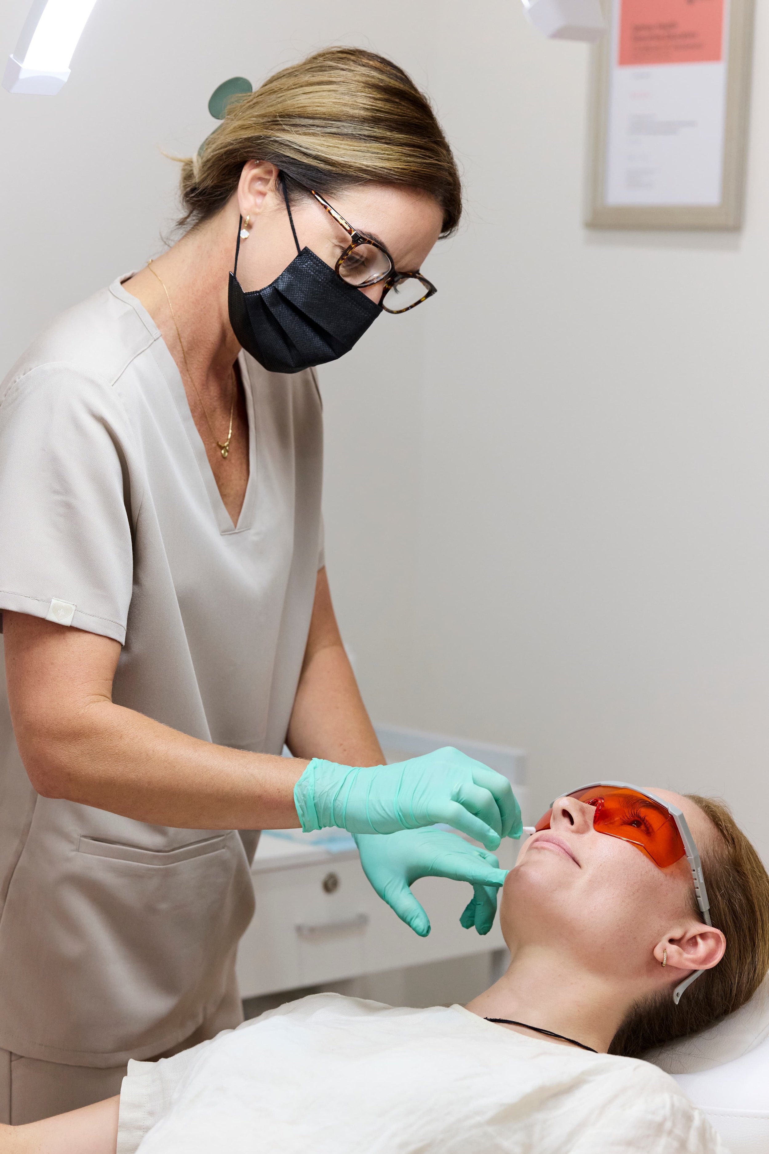 A dental professional wearing a black face mask, glasses, and green gloves administers a procedure to a patient lying in a dental chair, with protective orange eyewear.