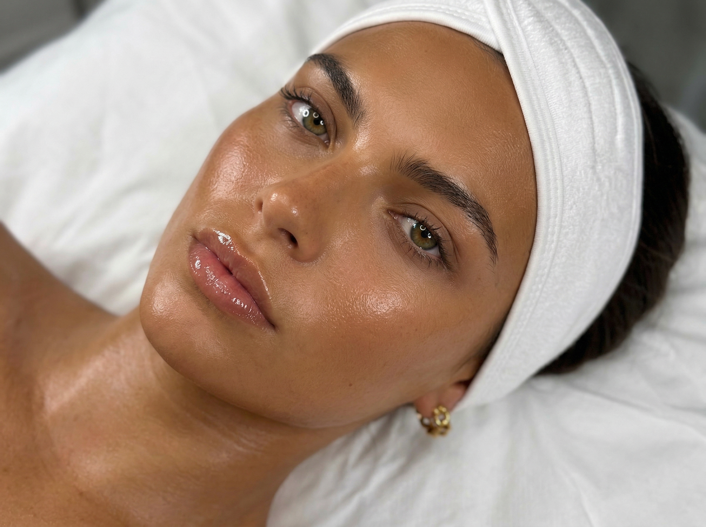 Close-up of a woman with clear, glowing skin lying on a white pillow, wearing a white headband and small gold earrings.