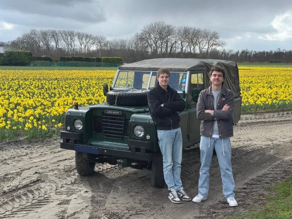 Twee jonge mannen staan naast een groene Land Rover op een zandweg, omringd door een veld met gele bloemen en bomen in de achtergrond onder een bewolkte hemel.