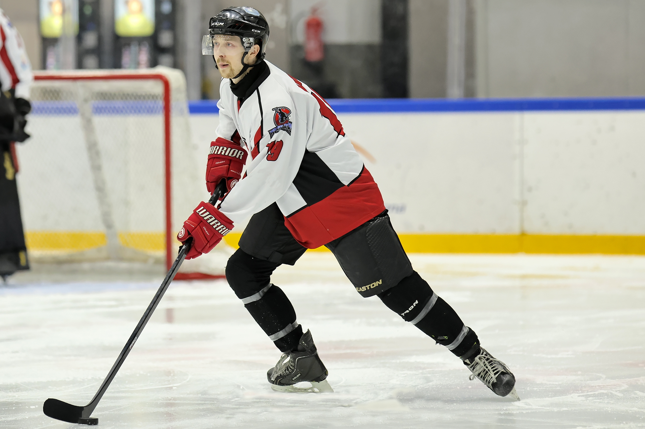 An ice hockey player wearing a white, black, and red jersey with a helmet, gloves, and skates on an ice rink, holding a hockey stick, with a hockey goal and other players in the background.