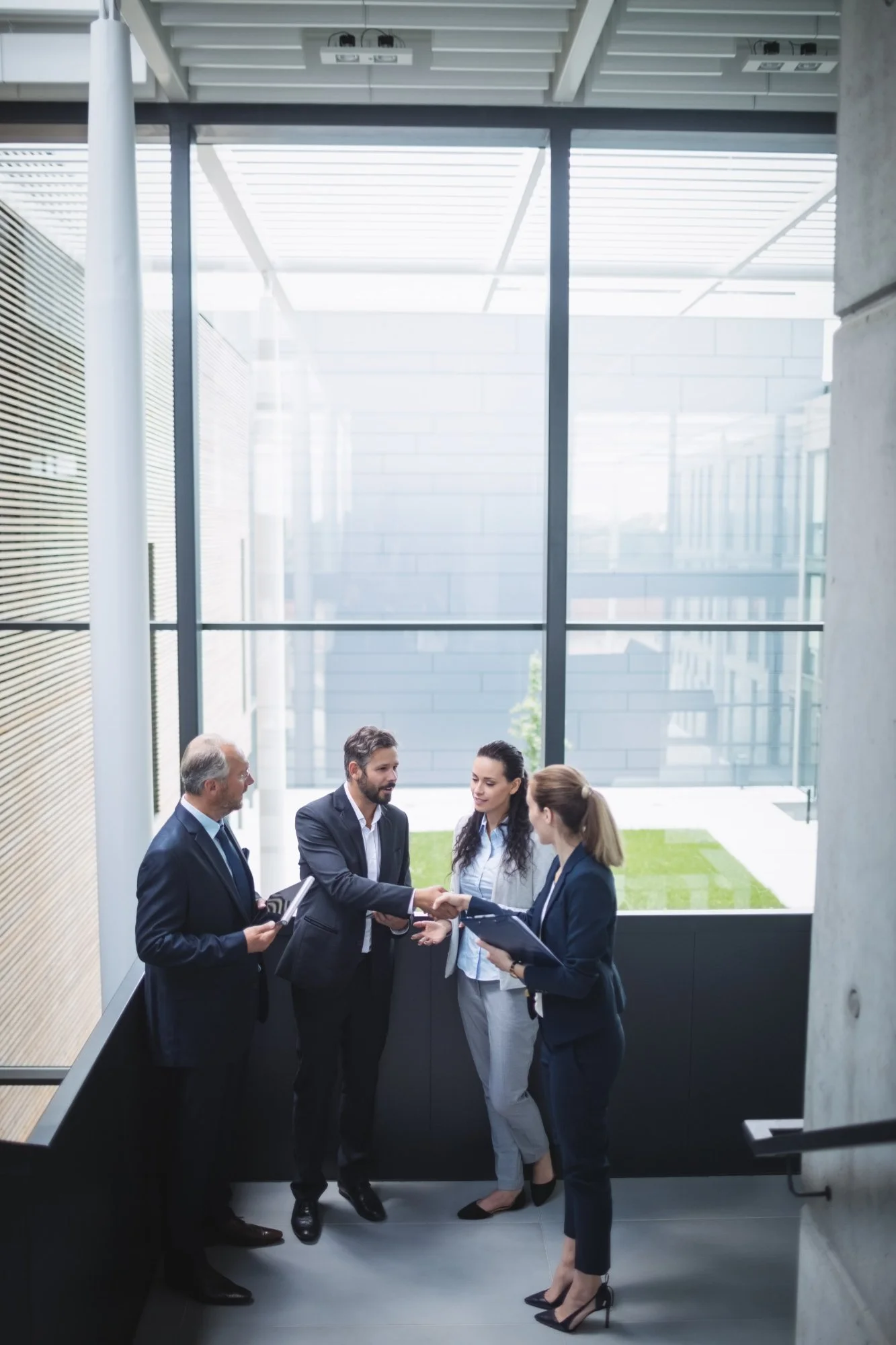 Four business professionals, two men and two women, socializing in a modern office lobby with large glass windows and a small outdoor green area.