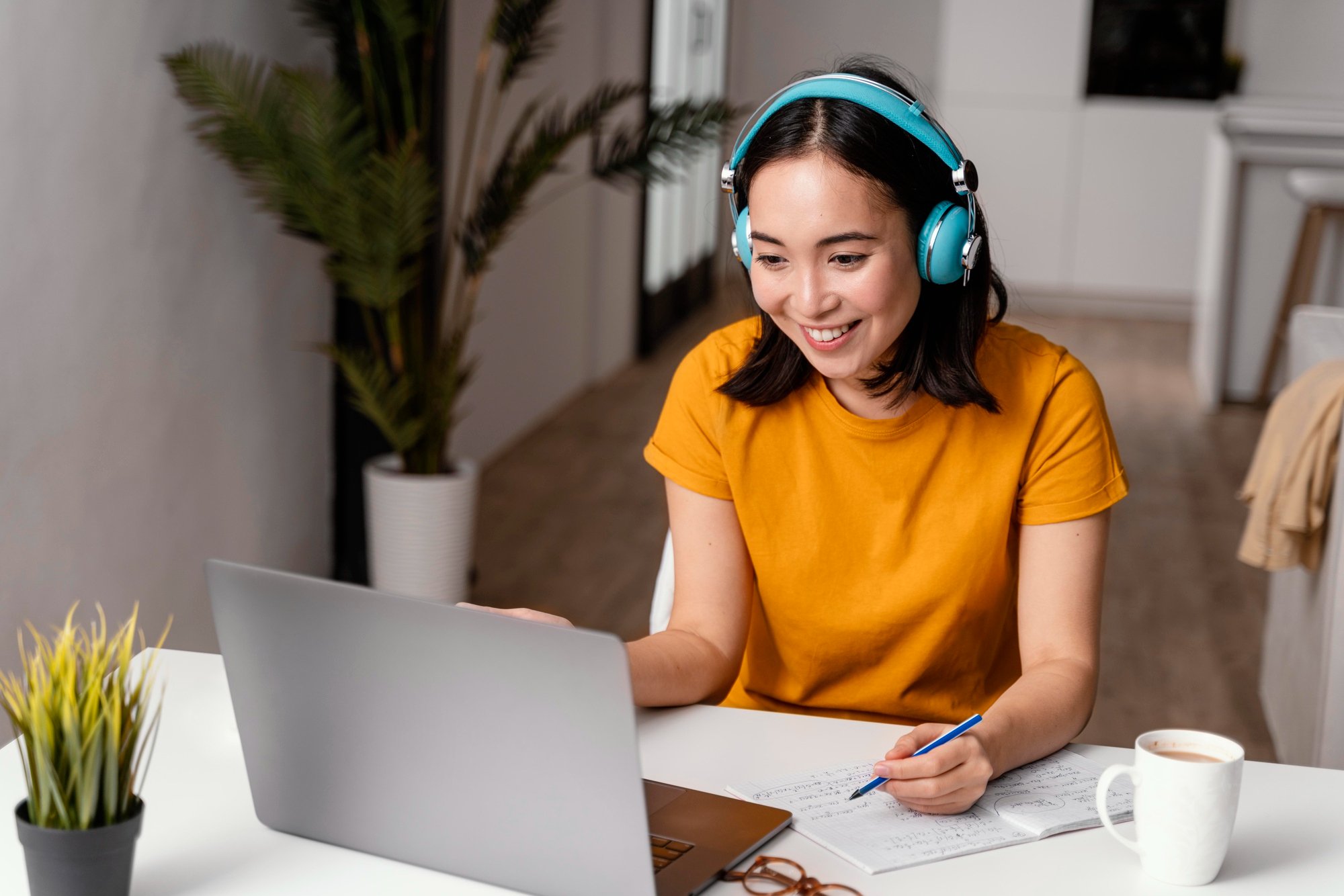 A woman smiling and wearing teal headphones while working at a desk with a laptop, notebook, and coffee mug