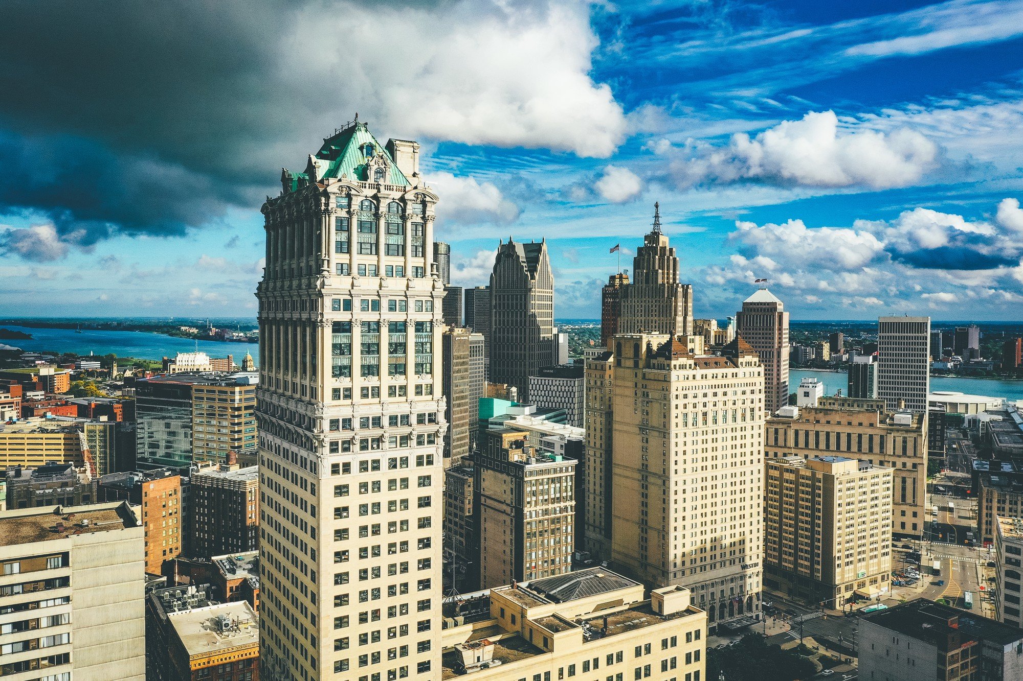 A city skyline with skyscrapers under a partly cloudy sky, with a river visible in the background.