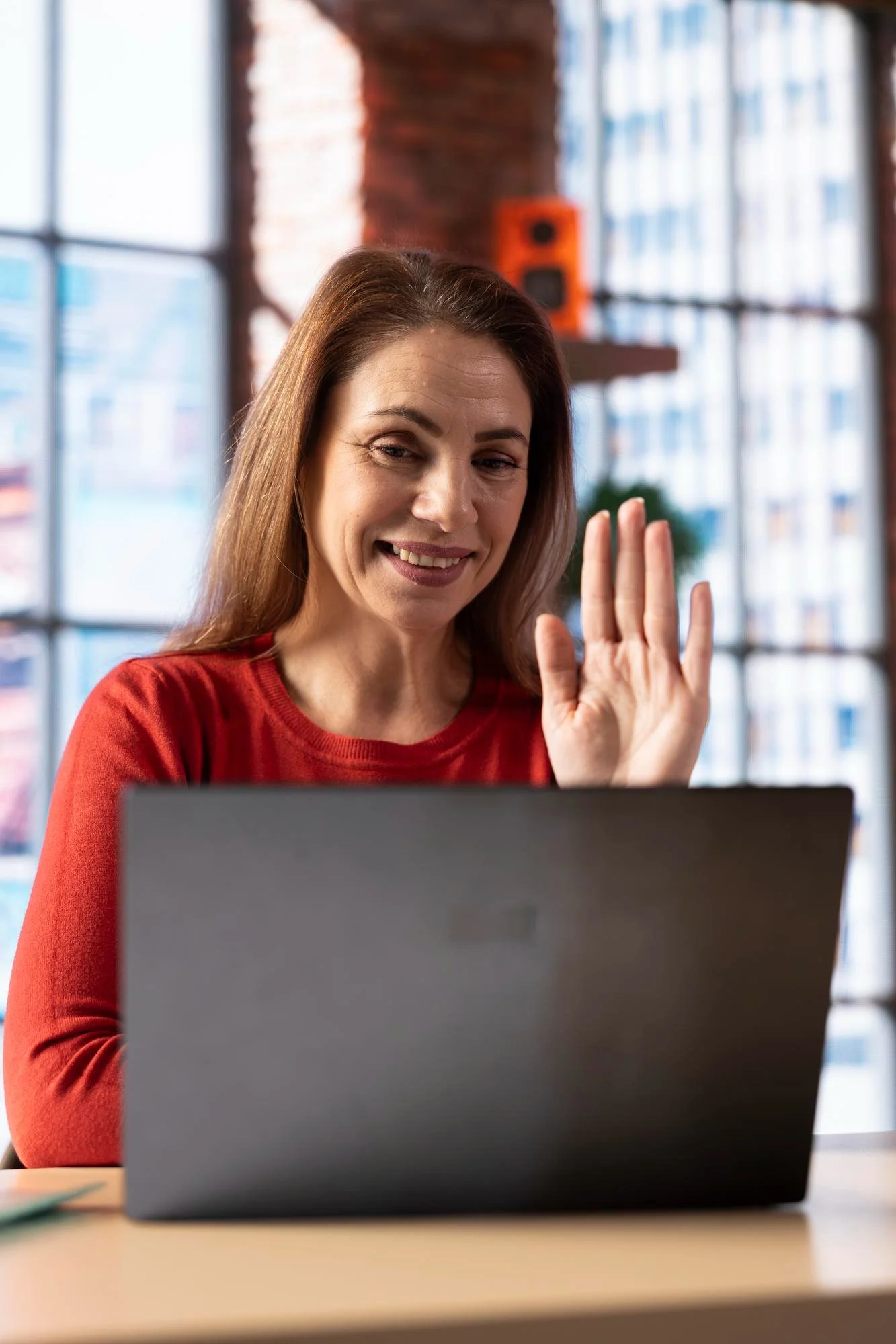 A woman with long brown hair smiling and waving at her laptop in a modern office with large windows and exposed brick.