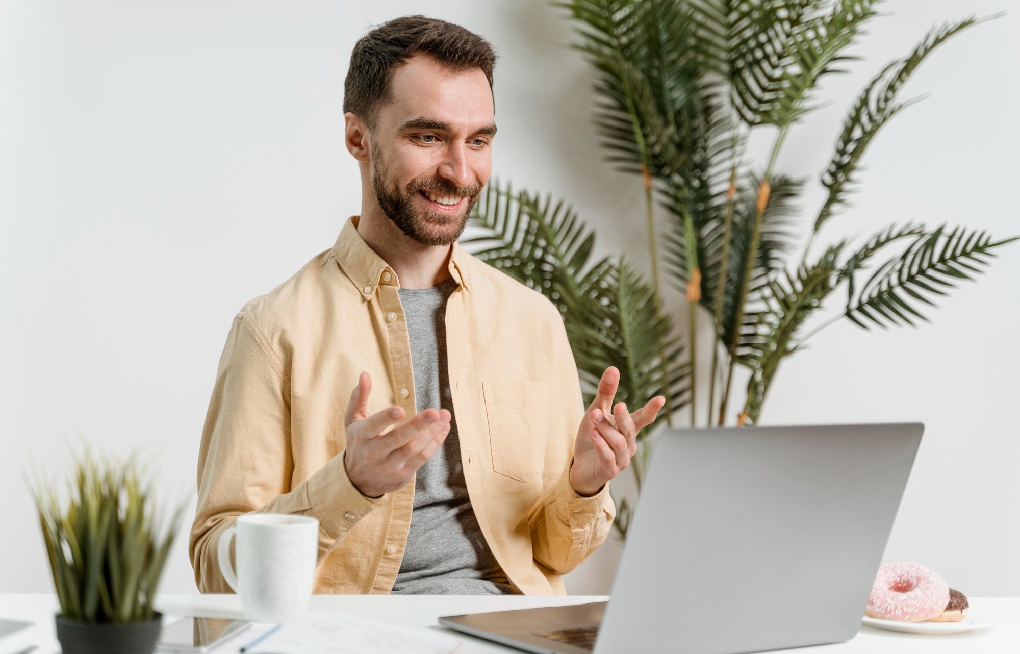 A man with a beard in a beige shirt smiling and gesturing while sitting in front of a laptop in a bright, modern room with green plants and a plate of donuts.