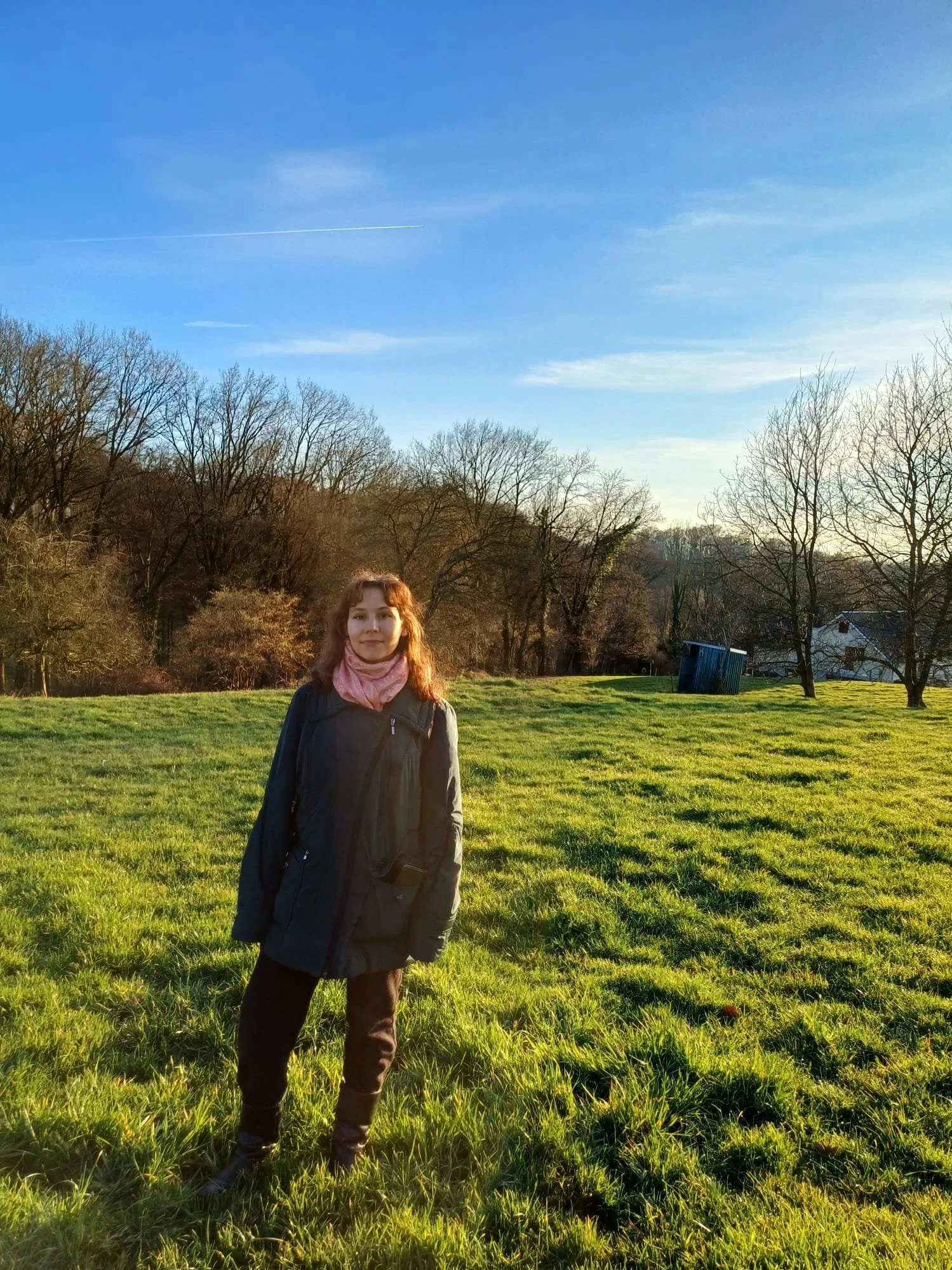 A woman standing in a grassy field with trees and houses in the background on a sunny day with a clear blue sky.