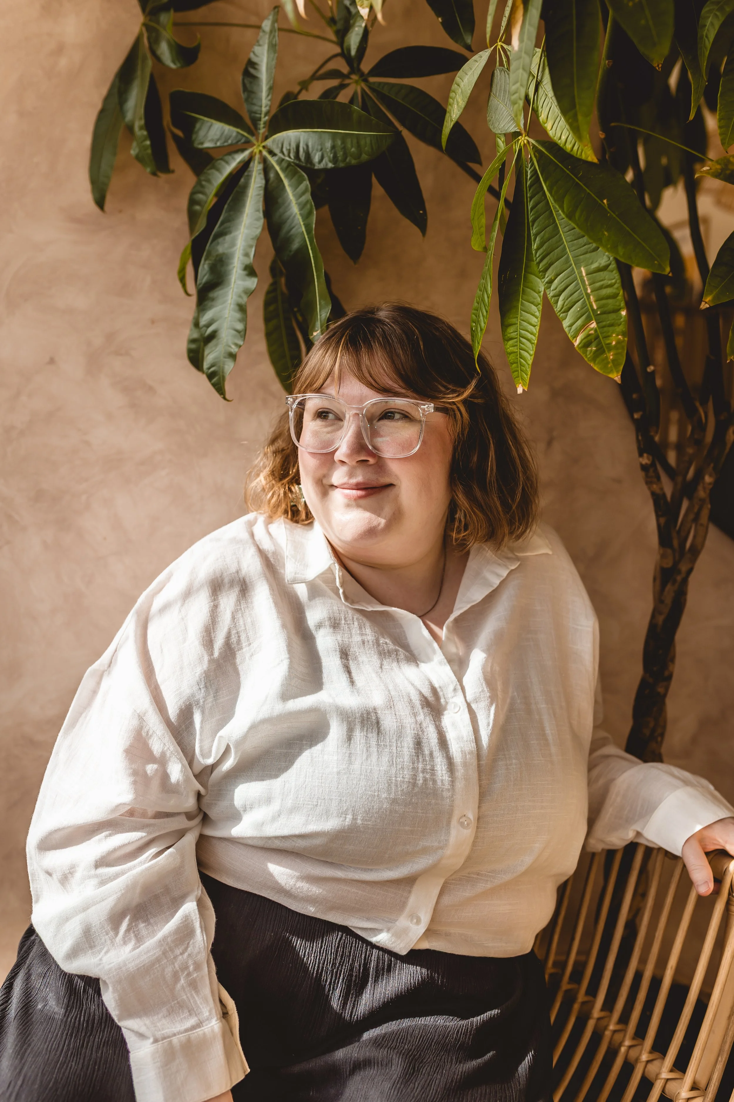 A woman with light skin and glasses sitting in front of a large leafy plant, wearing a white shirt and black skirt, smiling and looking to the side.