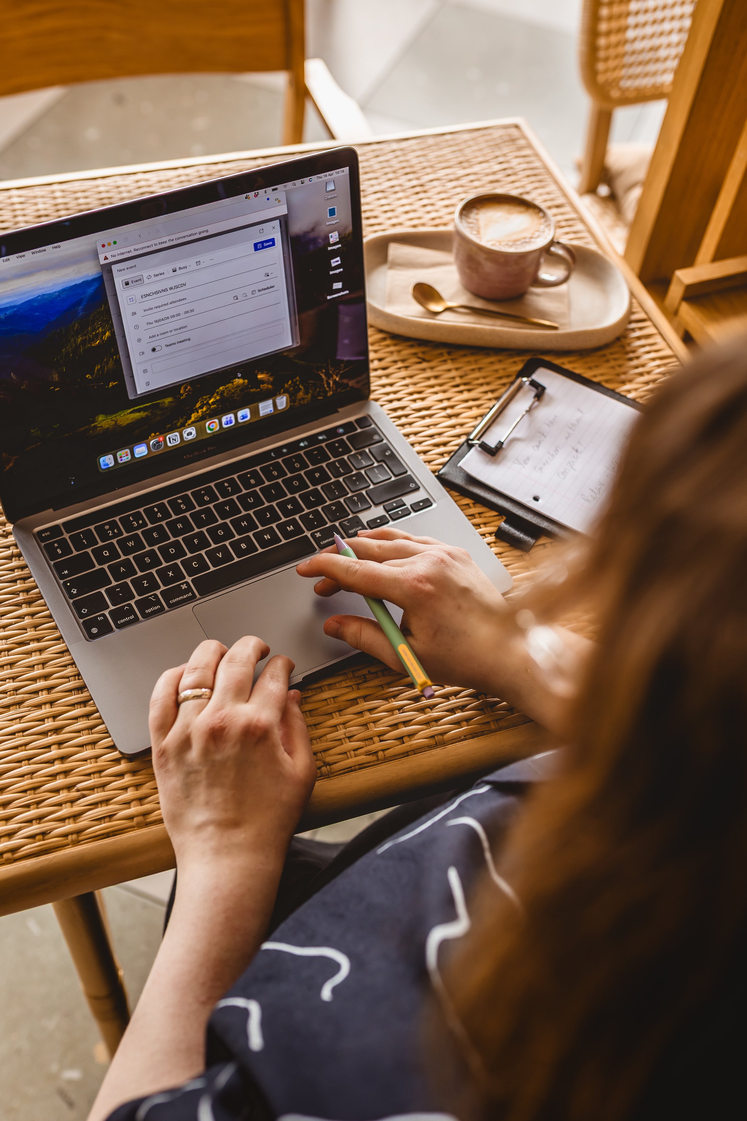 A person working on a MacBook laptop at a wicker table, with a notebook, pen, and a cup of coffee nearby.