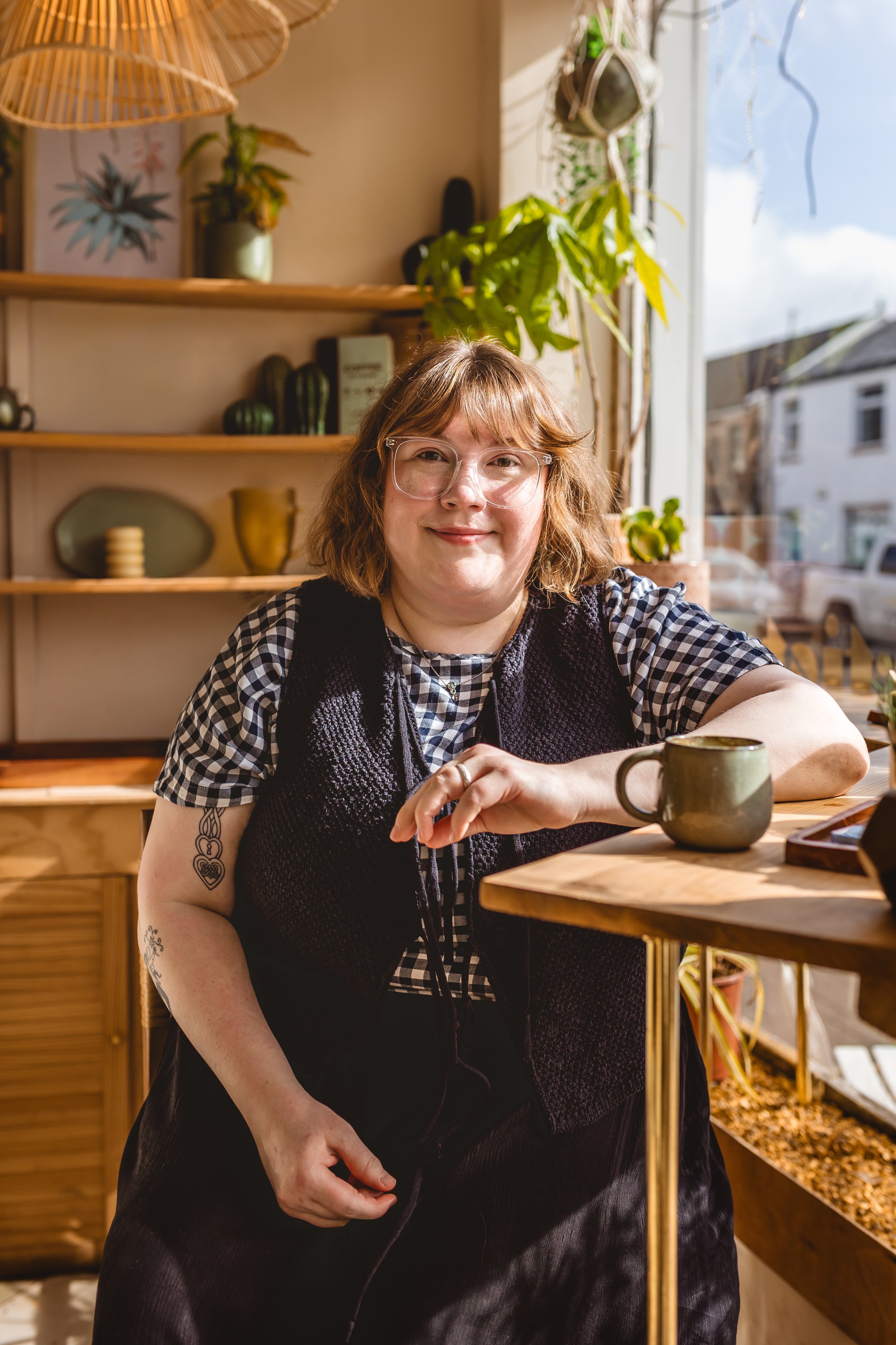 A woman with short, light brown hair and glasses sitting at a wooden table with a cup, in a cozy, well-lit space with shelves and plants.
