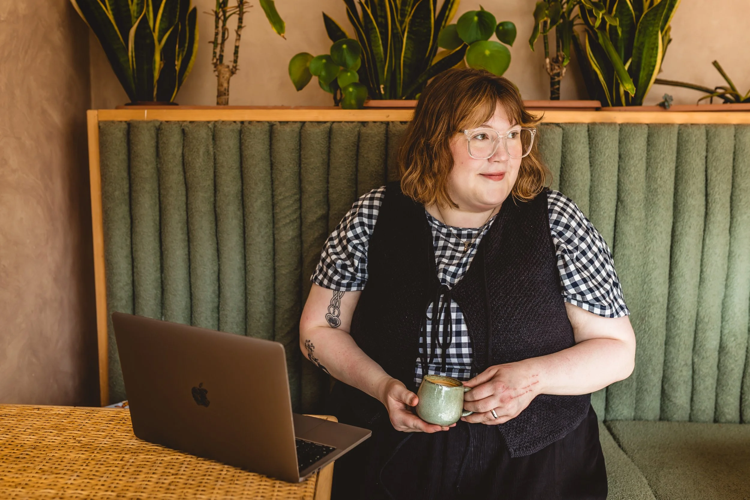 A woman with shoulder-length red hair and glasses holding a ceramic mug stands in front of a green upholstered booth with plants on a shelf behind her, next to a laptop on a woven table.